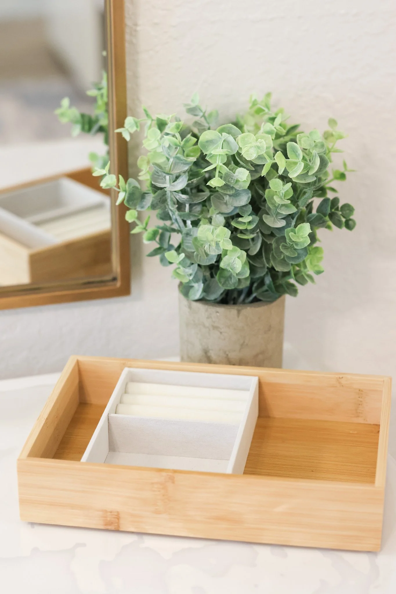 A decorative green artificial plant in a beige pot, placed next to a square wooden tray with smaller white trays inside, on a white surface near a wall and mirror.