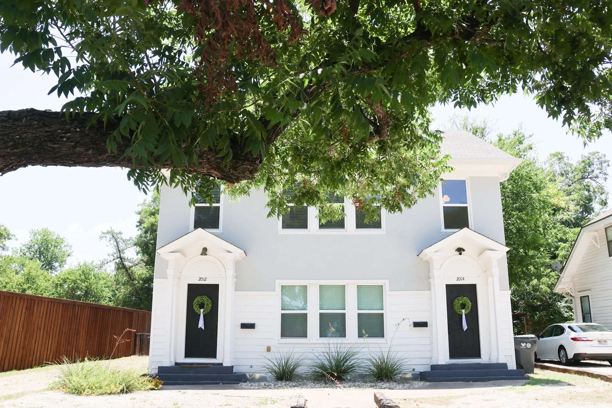 White two-story house with a black front door on the left and black door on the right, each with a green wreath, and a large tree with green foliage. The house has four windows on the upper level and four on the lower level. There is a wooden fence o