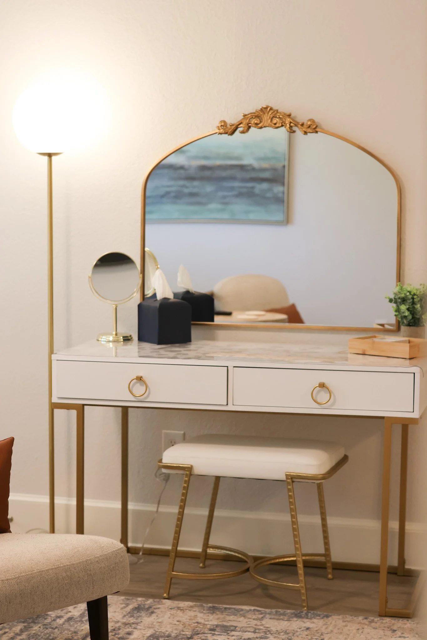 A white dresser with gold accents featuring a large ornate mirror, a small black tissue box, tissue papers, and decorative objects; gold leg stool with a cushioned seat is nearby, and a framed painting of the ocean hangs on the wall.