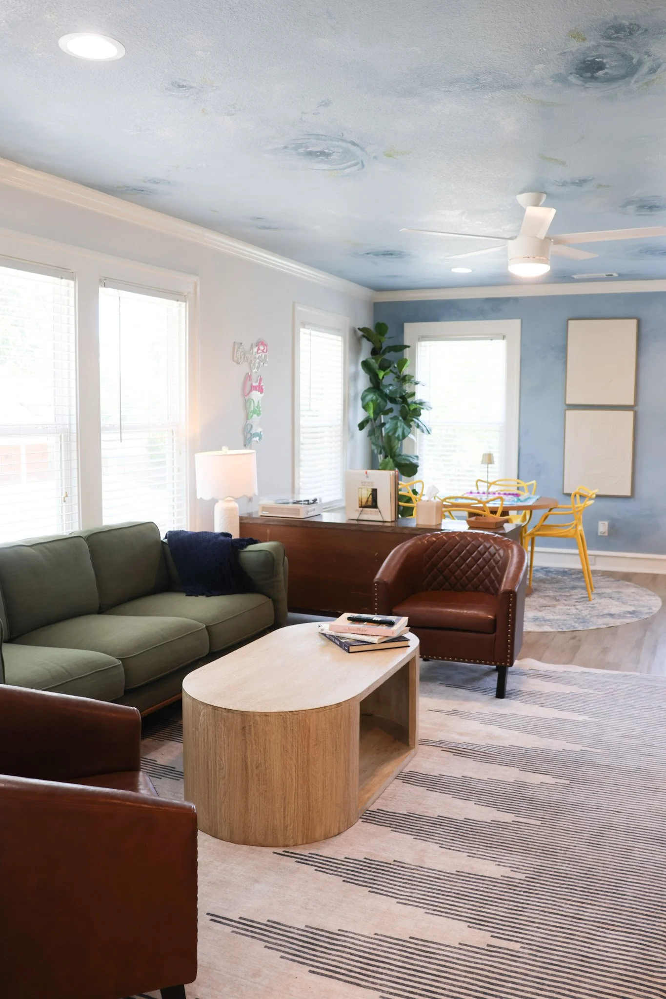 Living room with green sofa, brown leather chair, light wood coffee table, beige and gray carpet, tall plant, white ceiling fan, and large windows, with blue and white walls and decorative wall art.