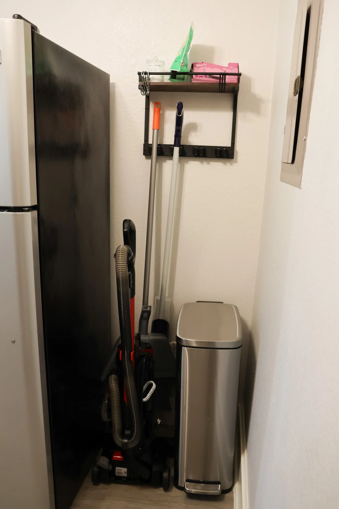 A small utility closet with cleaning supplies, cleaning tools, and a trash can, next to a refrigerator and a wall with an electrical panel.