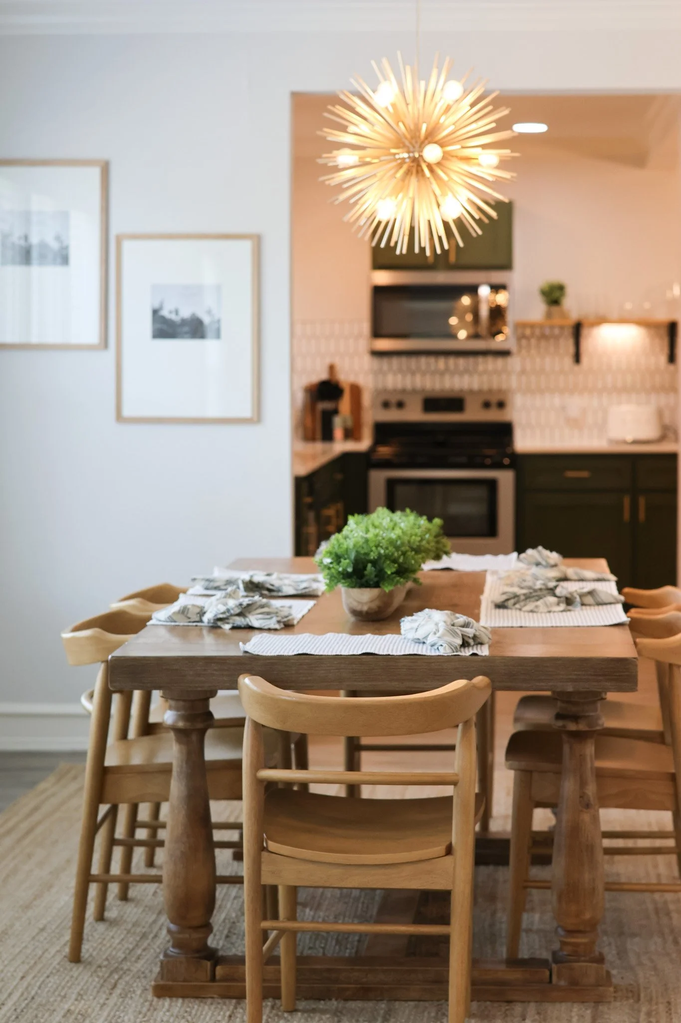 Wood dining table with nine matching wooden chairs and a potted plant centerpiece, situated in a kitchen area with hanging light fixture and two framed black-and-white photos on the wall.