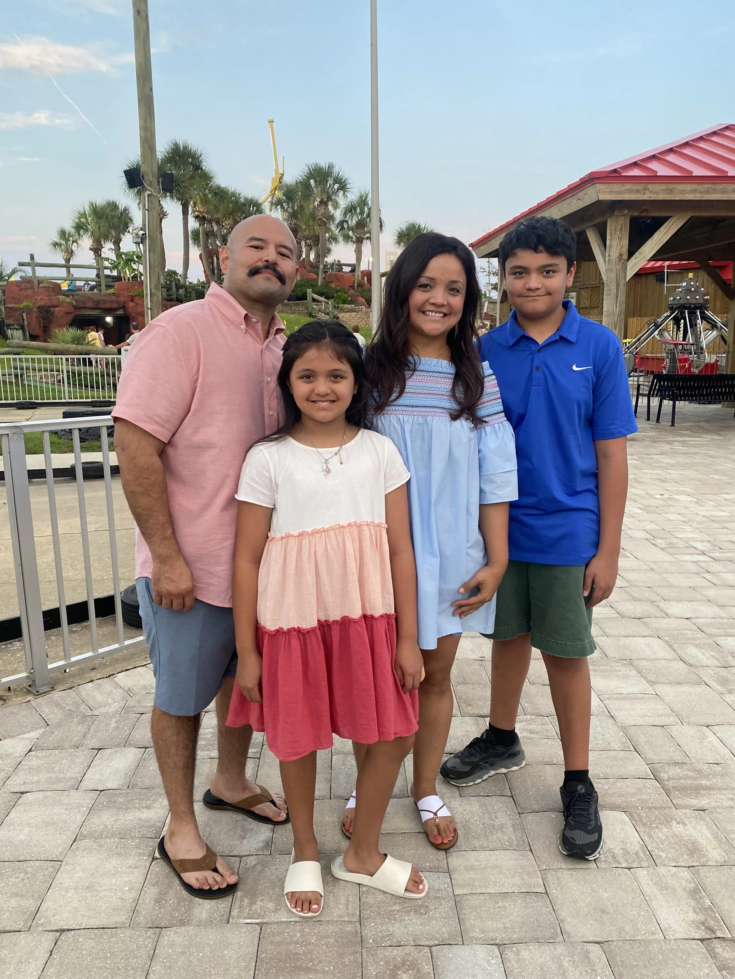 A group of four people standing outdoors on stone pavement, with palm trees, a red-roofed wooden structure, and amusement park rides in the background during daytime. The group includes a man with a mustache in a pink shirt and blue shorts, a young girl in a white and peach dress, a woman in a light blue dress, and a teenage boy in a blue Nike polo and dark green shorts, all smiling at the camera.