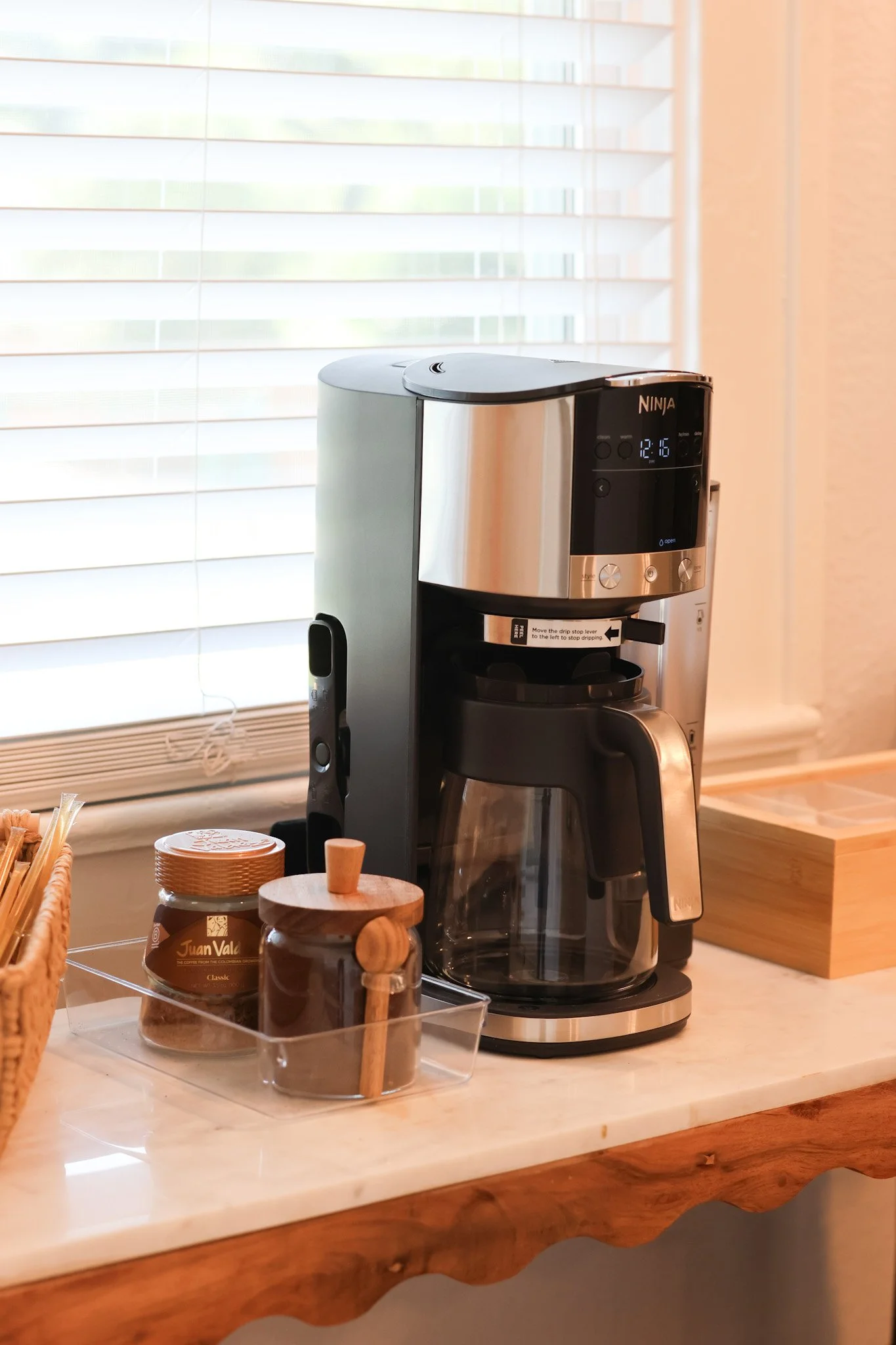 A coffee maker on a kitchen counter next to jars of sugar and spices, with a wooden knife block and other kitchen items nearby, positioned in front of a window with blinds.