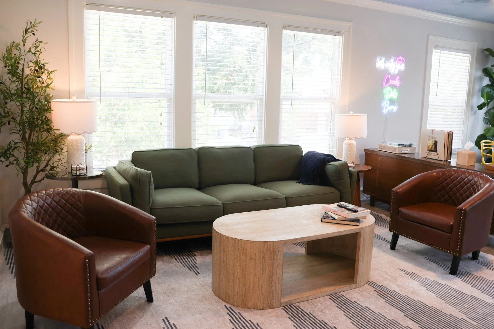 Living room with green sofa, two brown leather chairs, a wooden coffee table with books, large windows with white blinds, a tall plant, a neon sign on the wall that reads 'Hello, Craft My Brain' in colorful lights, and a side table with lamps.