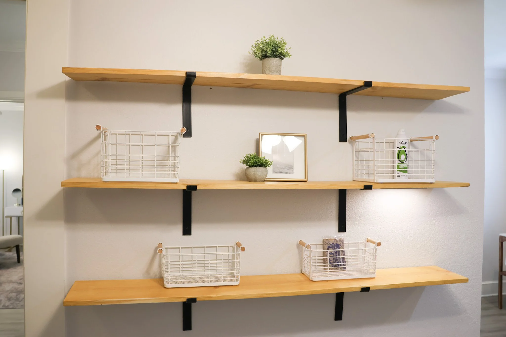 Wall-mounted wooden shelves with black brackets, holding small potted plants, picture frames, and storage baskets against a plain white wall.