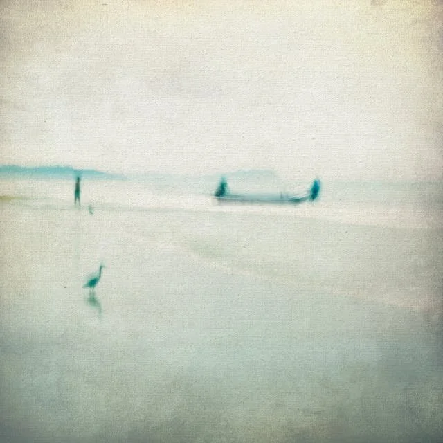 A blurry image of a person walking on a beach near the water, with a boat in the background and a bird perched on the sand in the foreground.