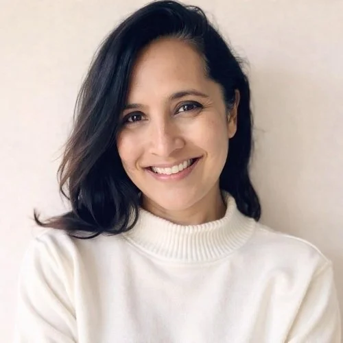 A woman smiling with black shoulder-length hair, wearing a white sweater, standing against a beige wall.