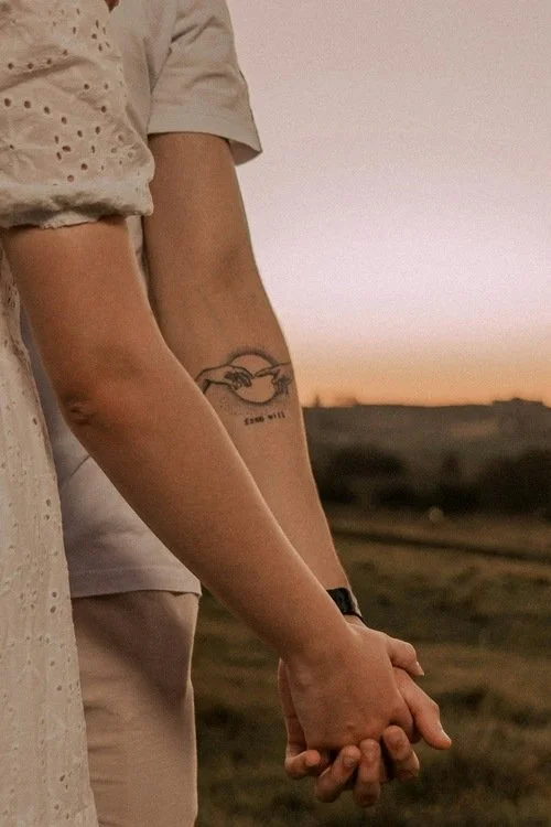 Two people holding hands outdoors during sunset, with one person's arm visible showing a tattoo of a bird and a phrase.
