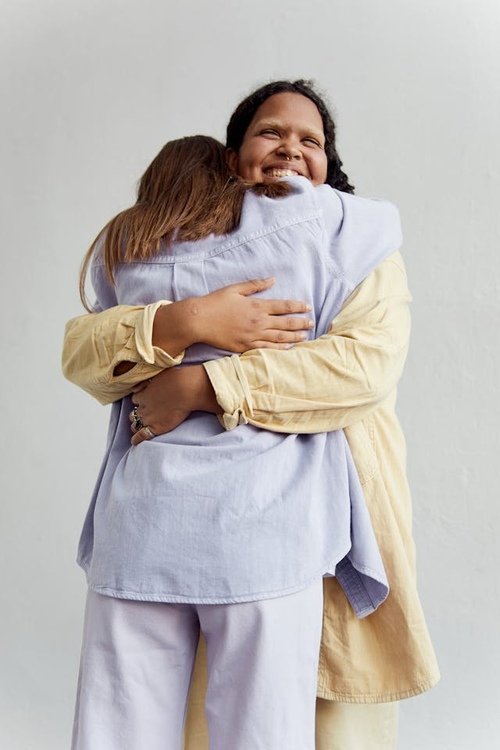 Two women hugging and smiling, one with darker skin and curly hair, the other with lighter skin and straight hair, against a plain white background.