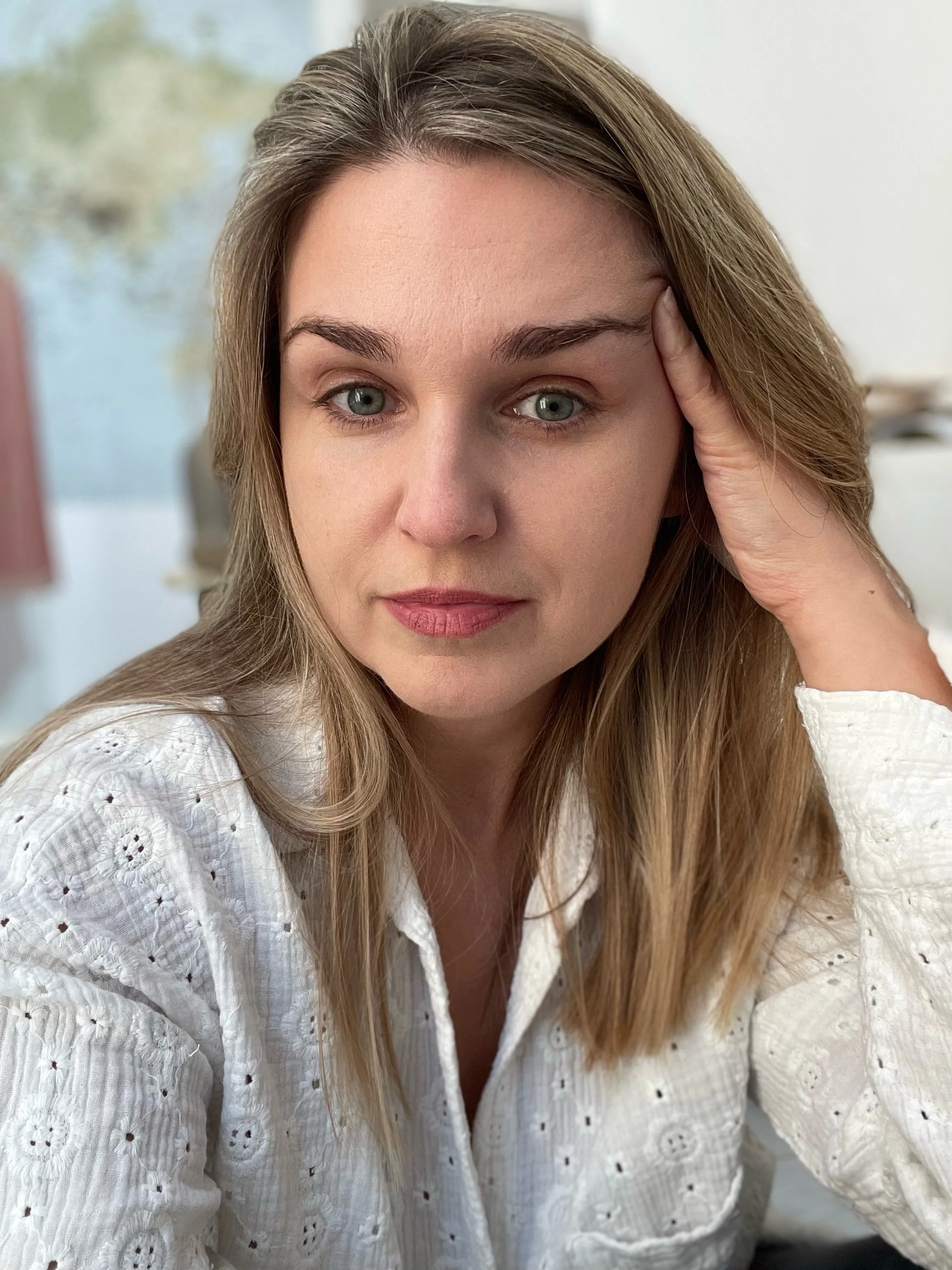 Close-up of a woman with blue eyes, light brown hair, wearing a white eyelet blouse, sitting indoors with a blurred background.