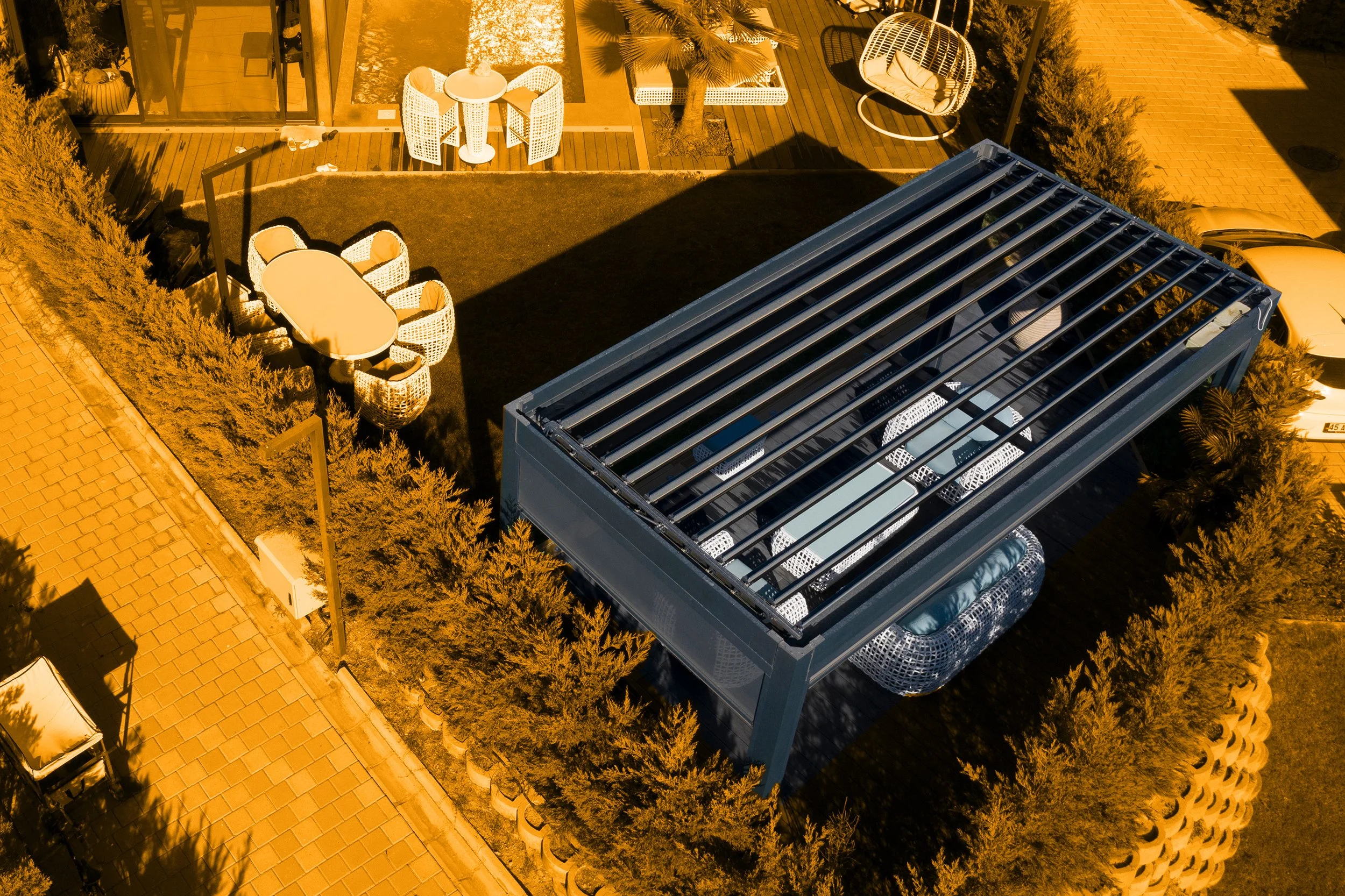 Aufnahme eines Gartenbereichs mit einem modernen blauen Carport, weißen Sitzmöbeln, einem runden weißen Tisch, umgeben von Büschen, einer Terrasse mit Sessel und Sonnenliegen, und einer gepflasterten Auffahrt.