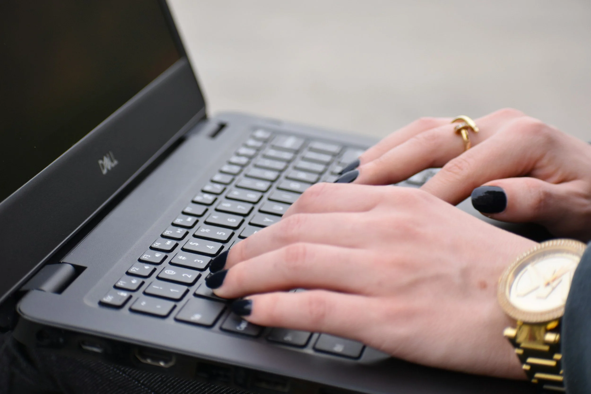 Close-up of hands typing on a Dell laptop keyboard, with black nail polish, wearing a gold wristwatch and a ring.