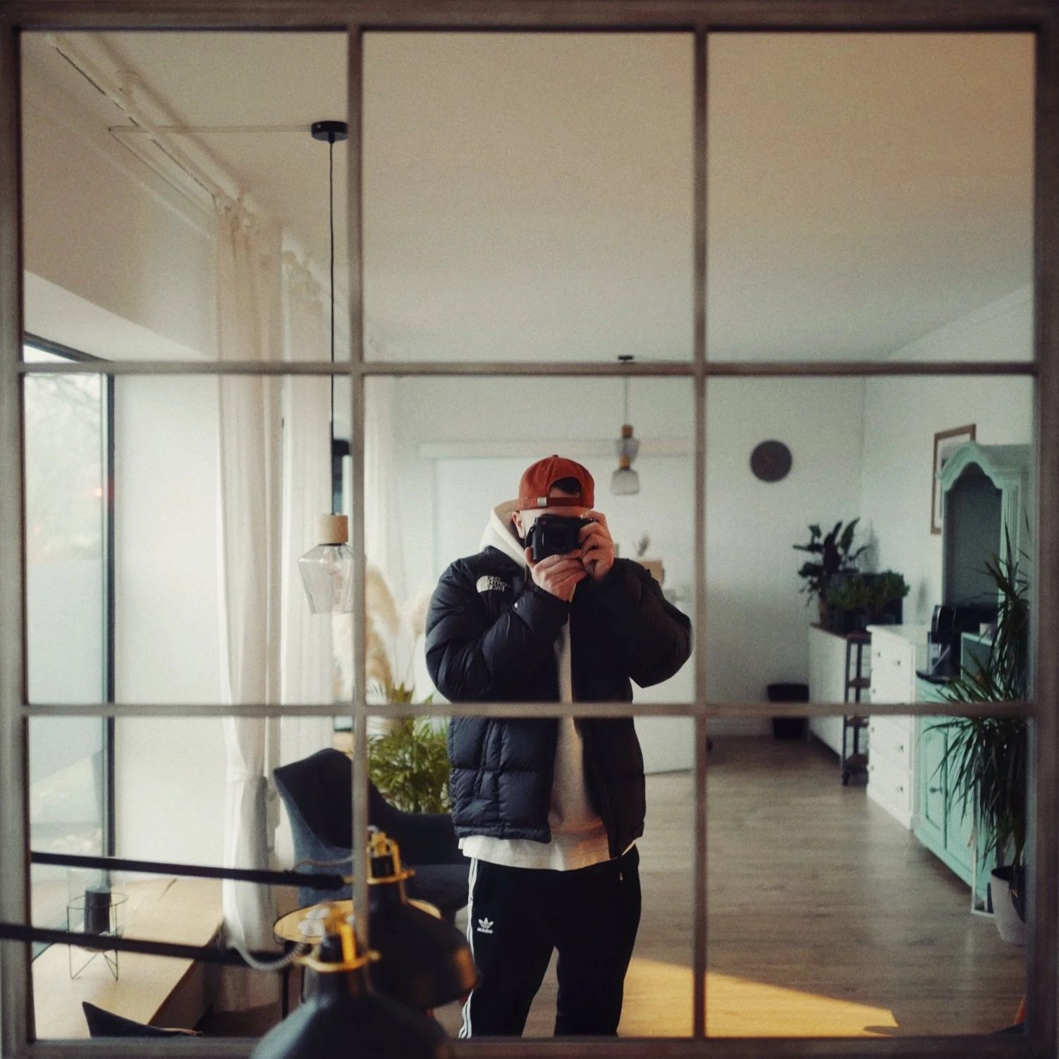 Person nimmt Selfie vor einem Spiegel in einem Wohnzimmer, trägt rote Kappe, schwarze Jacke, weiße Shirt, schwarze Jogginghose, im Hintergrund Pflanzen, Fernseher und Möbel.