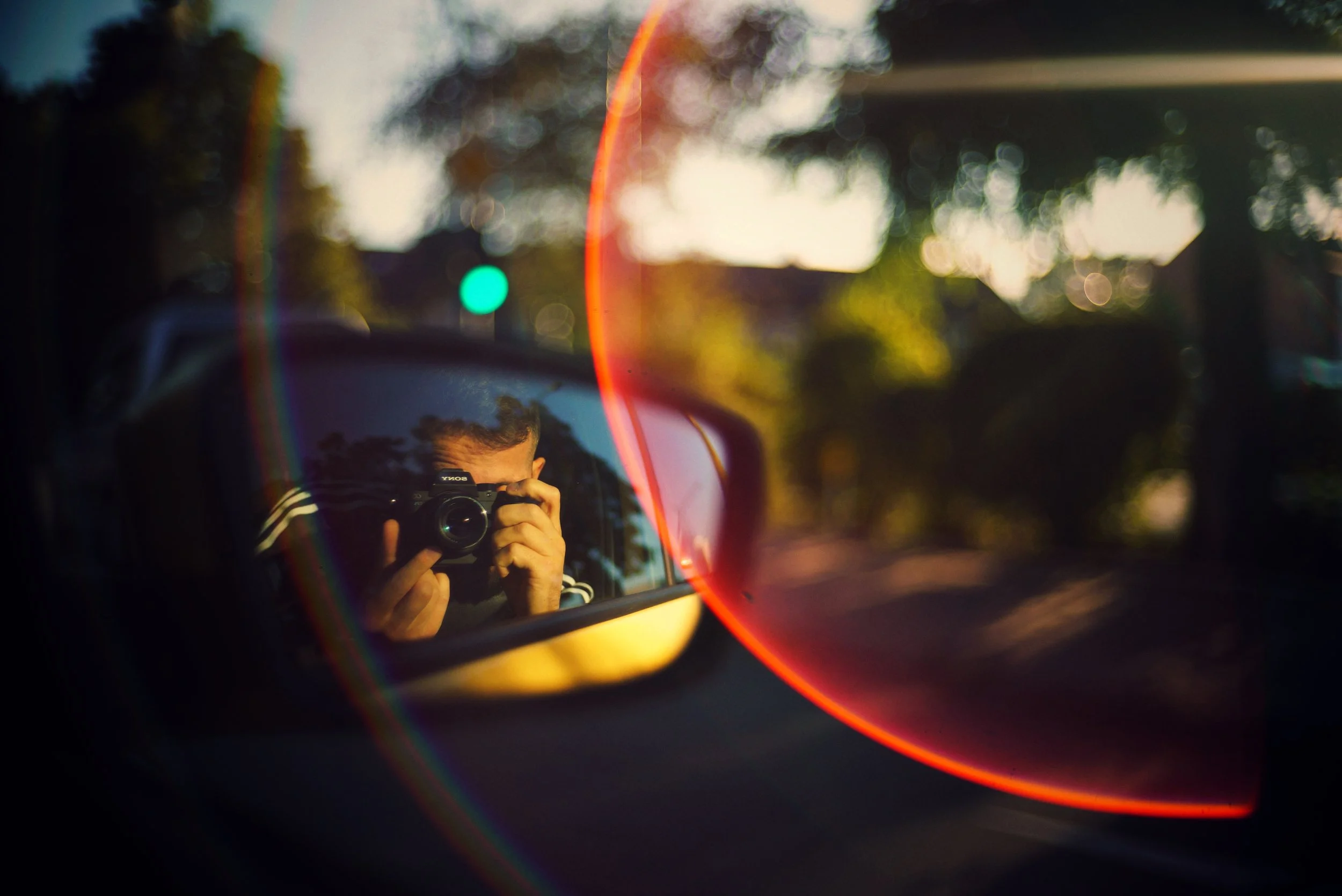 Das Bild zeigt den Spiegel eines Autos, in dem eine Person mit einer Kamera zu sehen ist, die den Spiegel fotografiert. Im Hintergrund sind Bäume und eine Straße bei Sonnenuntergang zu erkennen.