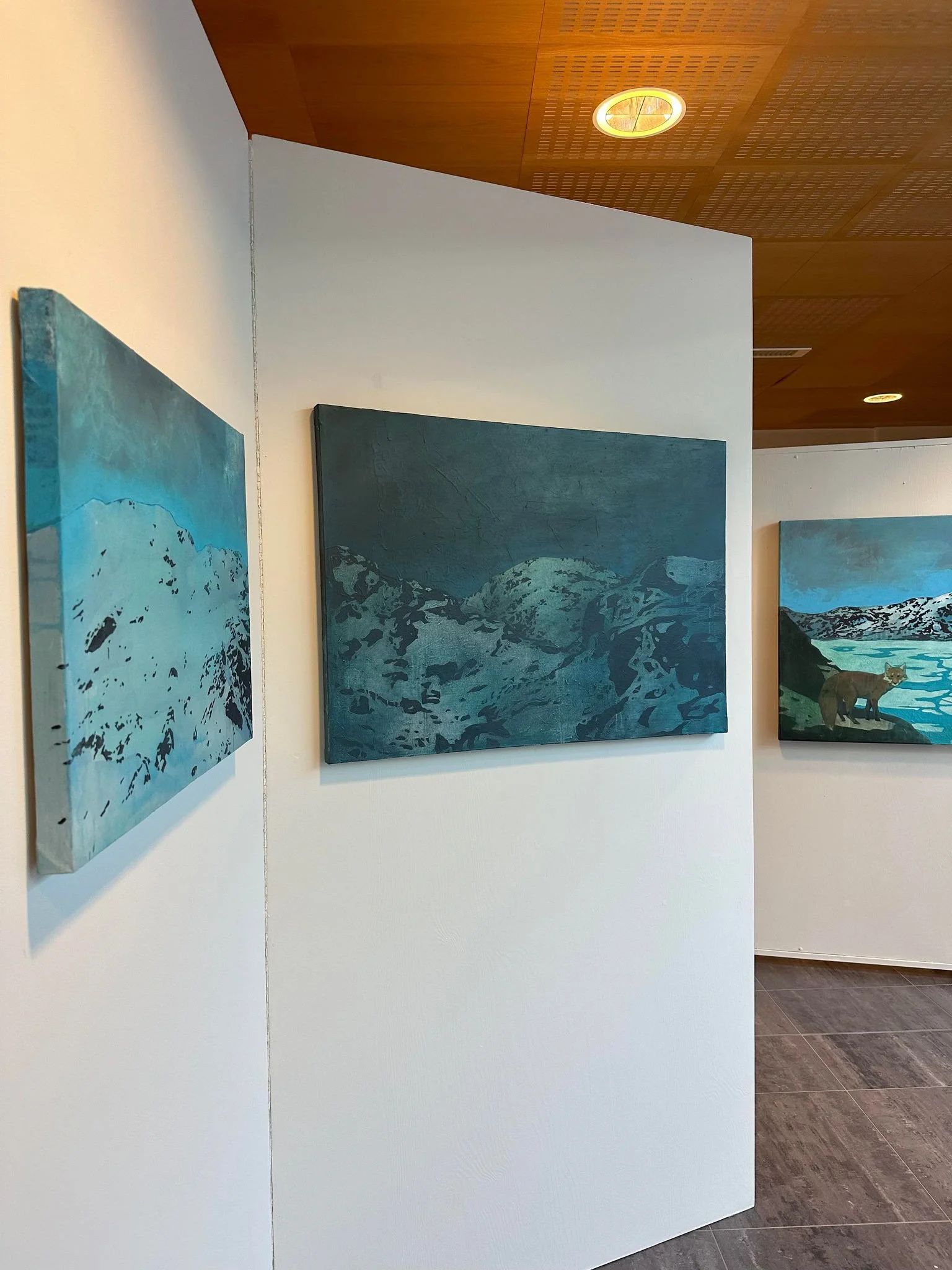 Bilde av tre malerier med fjellandskap i blått, hengt på en hvit vegg i et galleri, med taklampe i treverk og gulv fliser.