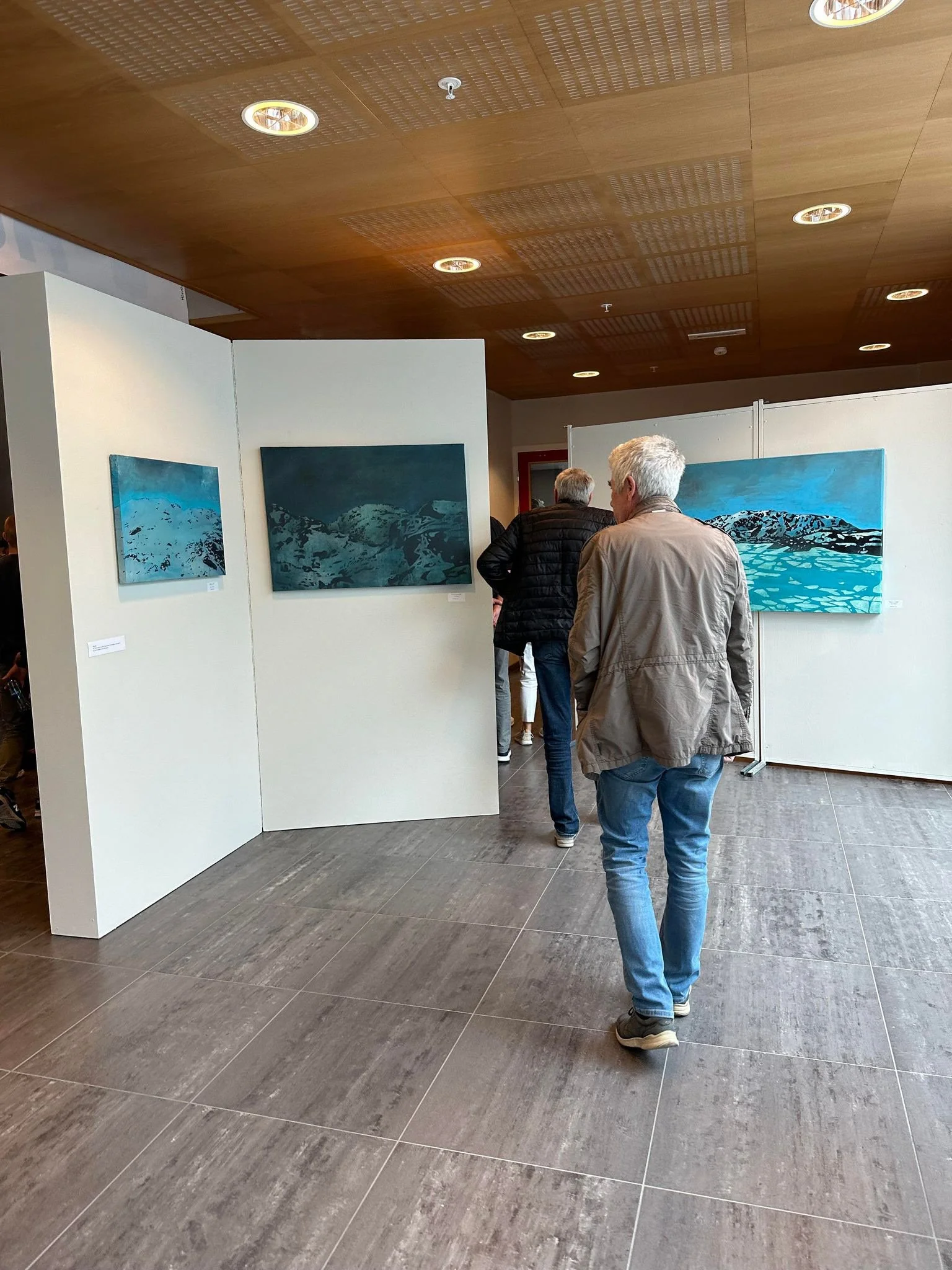 To personer ser på kunstverk i et galleri, med tre kunstverk på veggen, og har grå hår og beige/lyse jakker.
