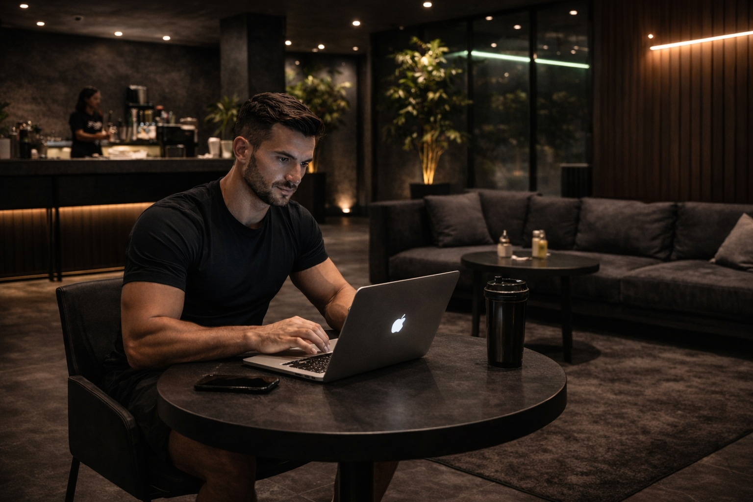 A man sitting at a round table using a silver MacBook in a dimly lit lounge or café with dark interior, couches, a coffee table, and a bar in the background.