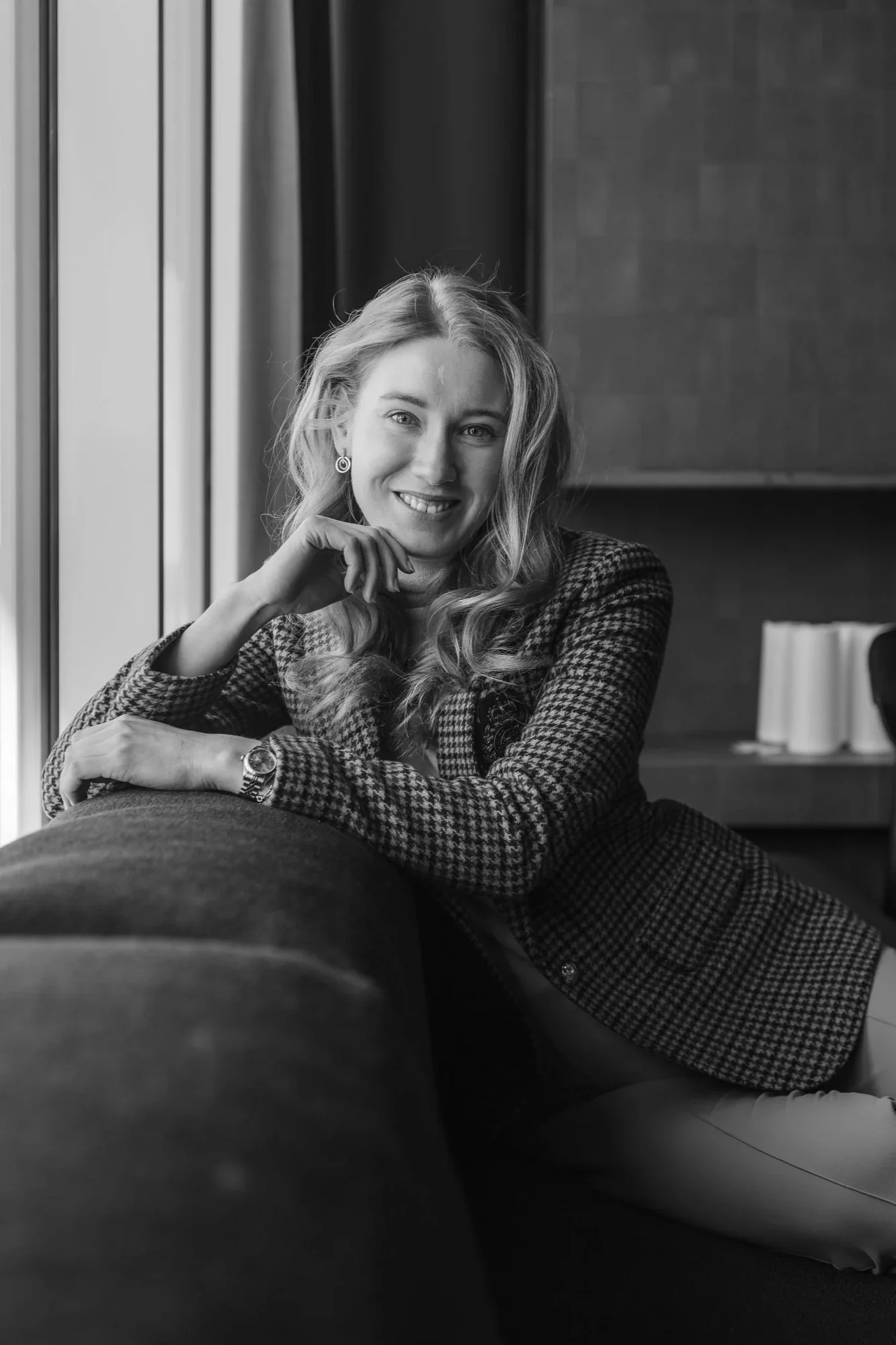 A woman with wavy hair smiling, sitting on a couch near a window, in a black and white photo. Henkilökuvaus lokaatiossa. Kuvauspaikka Valo Hotel & Work. Henkilöstökuvaus.