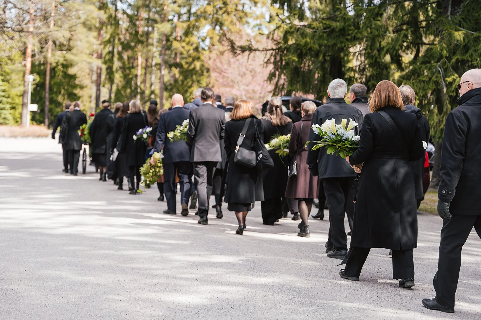 Saattoväki kävelemässä hautapaikalle arkun hautaan laskua varten. Mukanaan kukkalaitteet. Hautajaisten valokuvaus.