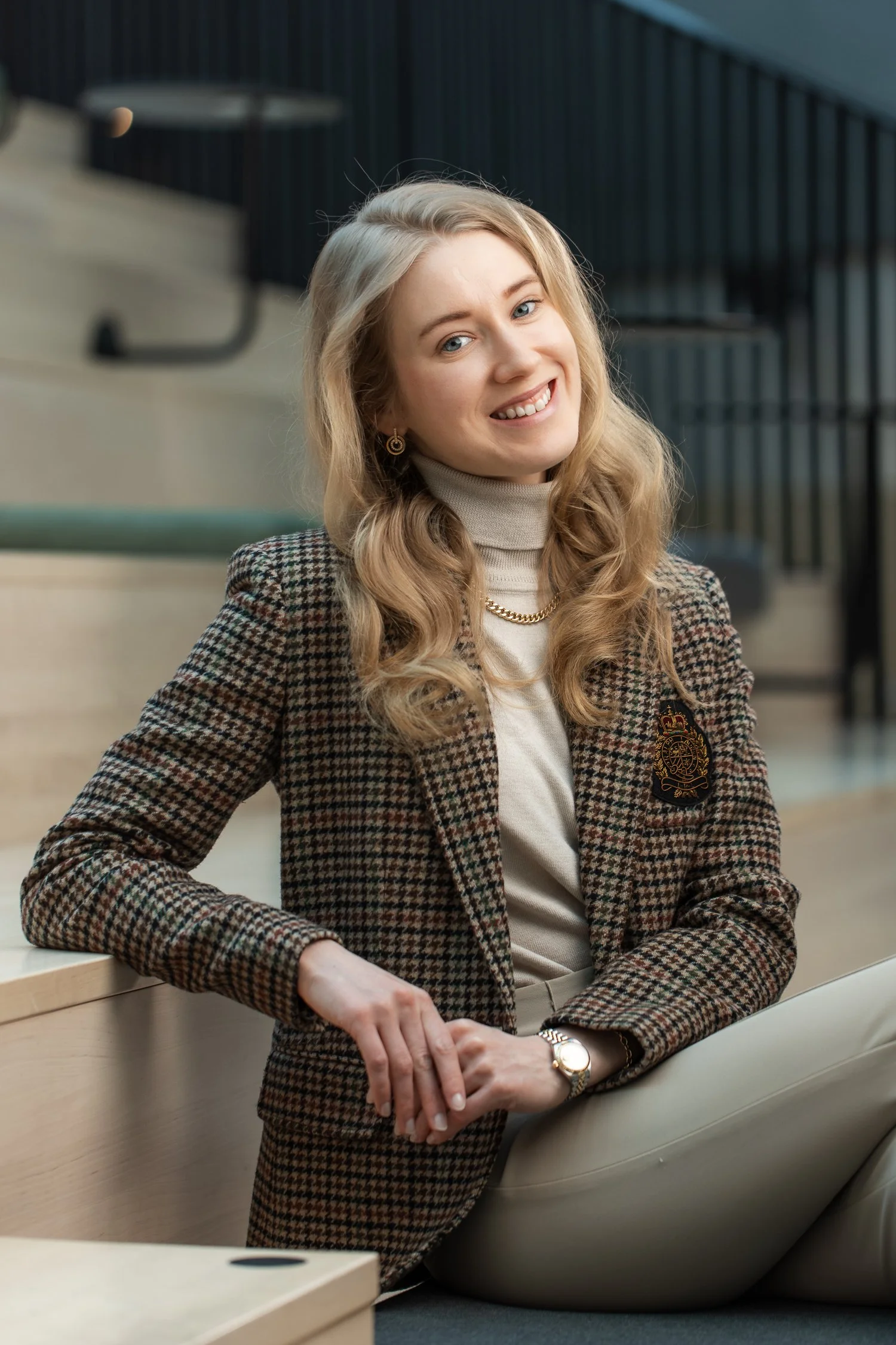 A woman with long curly blonde hair smiling, sitting on the floor in an indoor setting wearing a houndstooth blazer with a crest, a turtleneck, beige pants, and jewelry. Henkilökuvaus lokaatiossa. Kuvauspaikka Valo Hotel & Work. Henkilöstökuvaus.