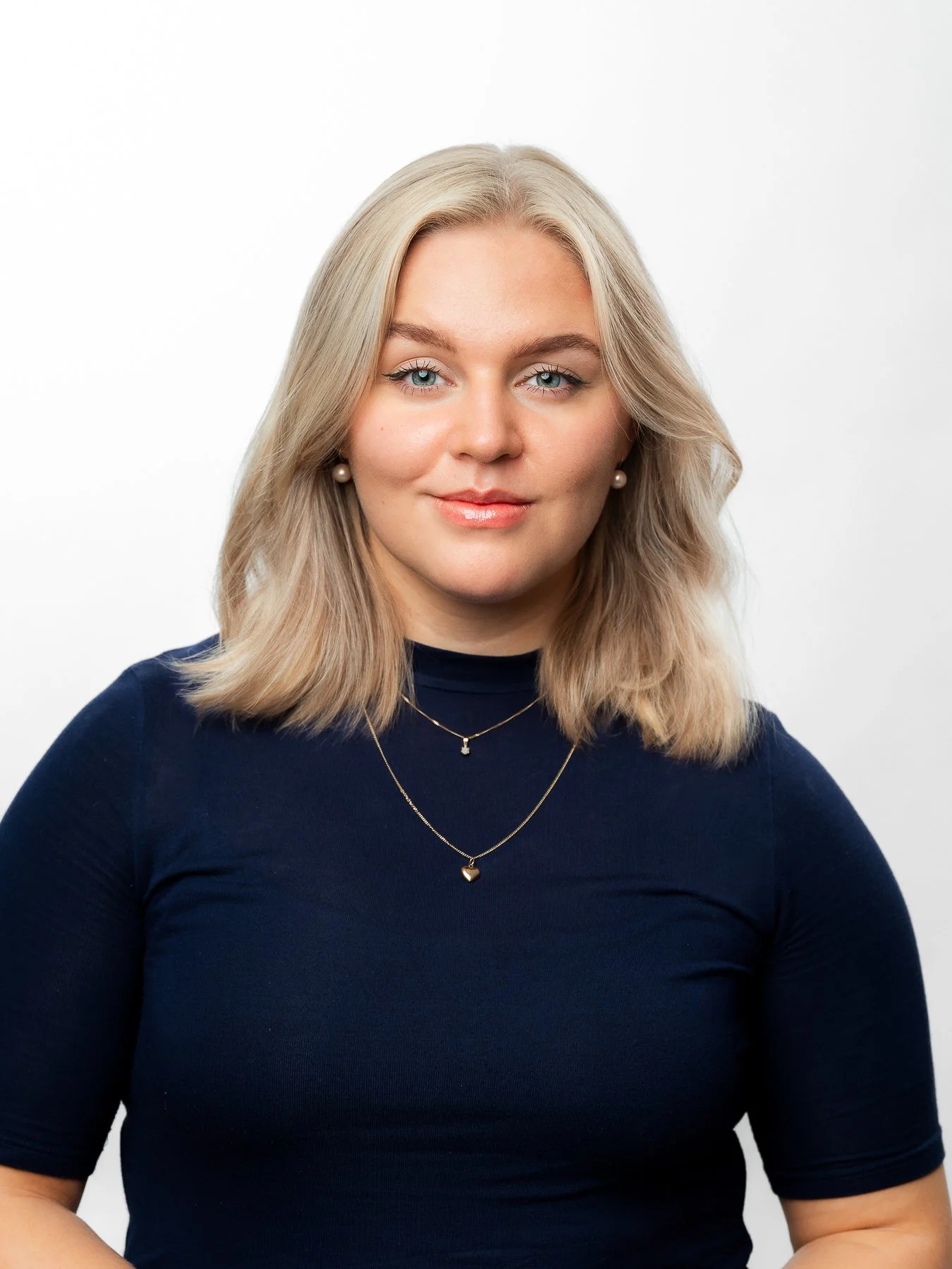 Portrait of a young woman with blonde hair, wearing a navy blue top, pearl earrings, and layered gold necklaces, smiling slightly against a plain white background. Yritysten henkilöstö valokuvaus. Henkilöstökuvaus studiossa. 