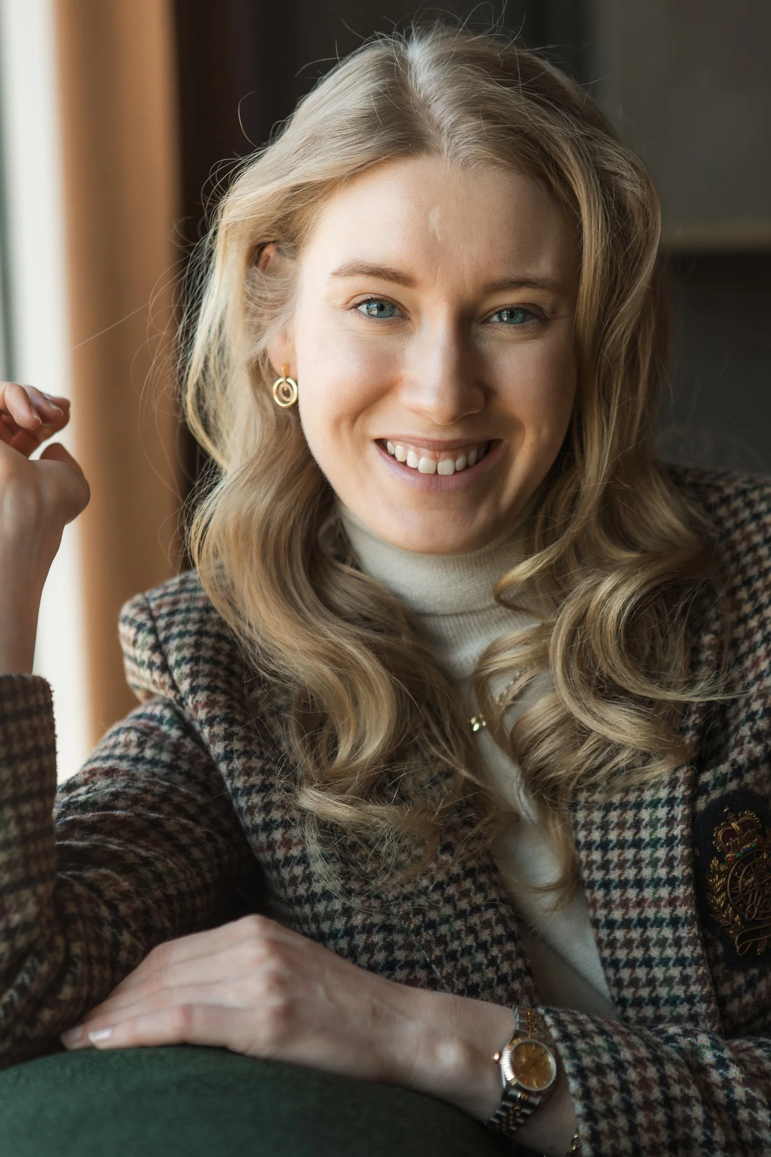 A young woman with blonde hair styled in loose waves, smiling at the camera, wearing a houndstooth blazer, white turtleneck, sitting indoors near a window. Henkilökuvaus lokaatiossa. Kuvauspaikka Valo Hotel & Work. Henkilöstökuvaus.