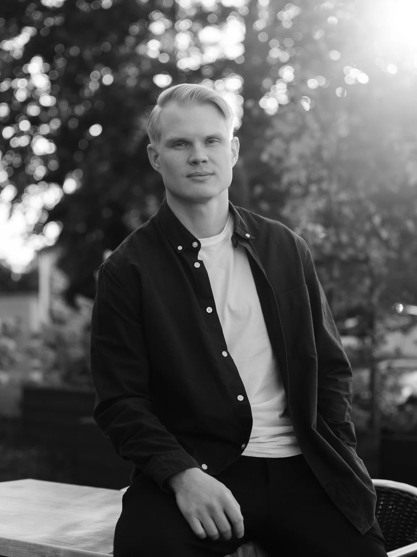A young man with blonde hair wearing a black button-up shirt and white t-shirt, sitting outdoors during sunset, with trees and sunlight in the background. Henkilökuvaus miljöössä.