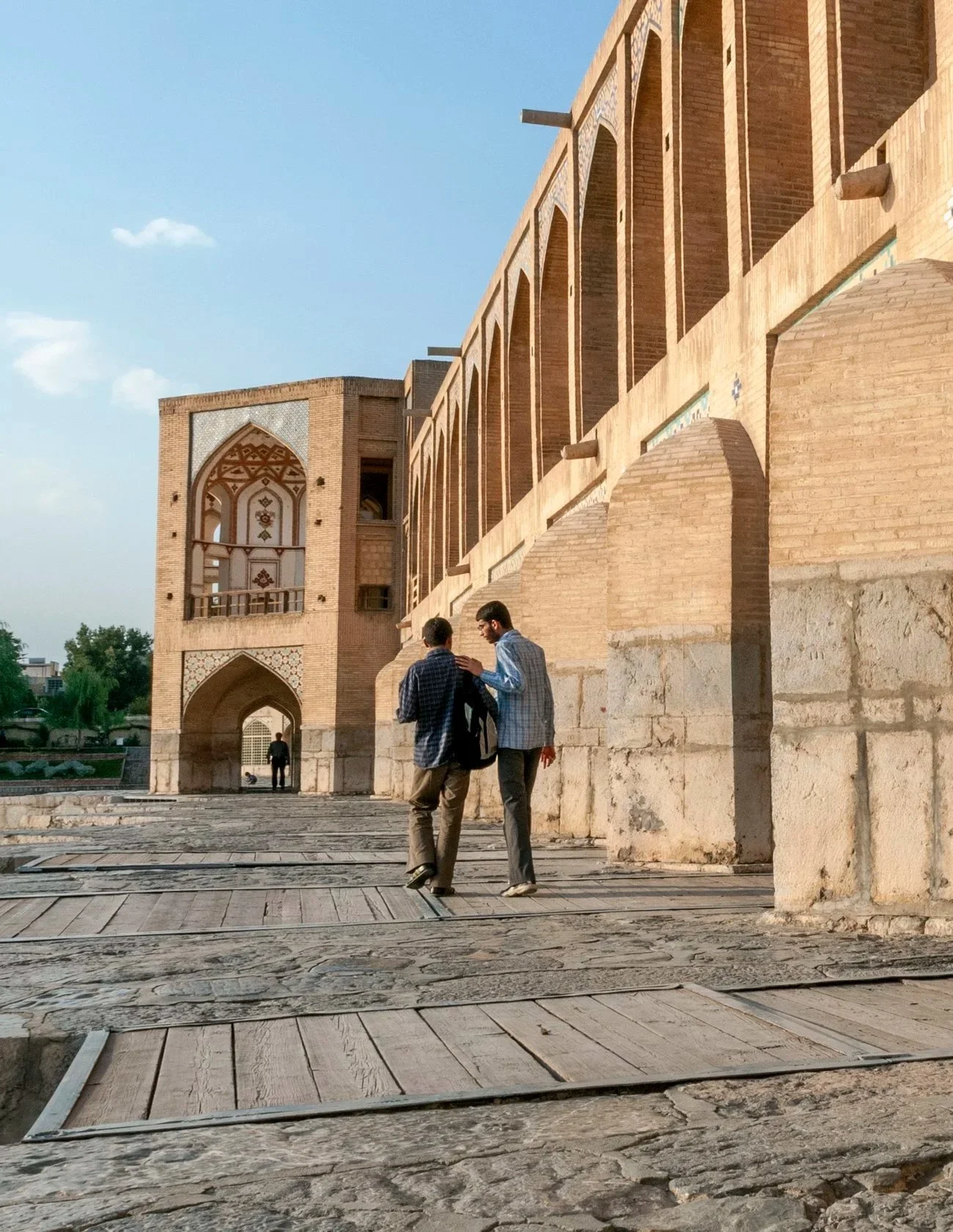 Two young men walking and talking near an ancient brick building with arched windows and decorative patterns. The scene is outdoors on a sunny day, with cobblestone and wooden plank pavement.