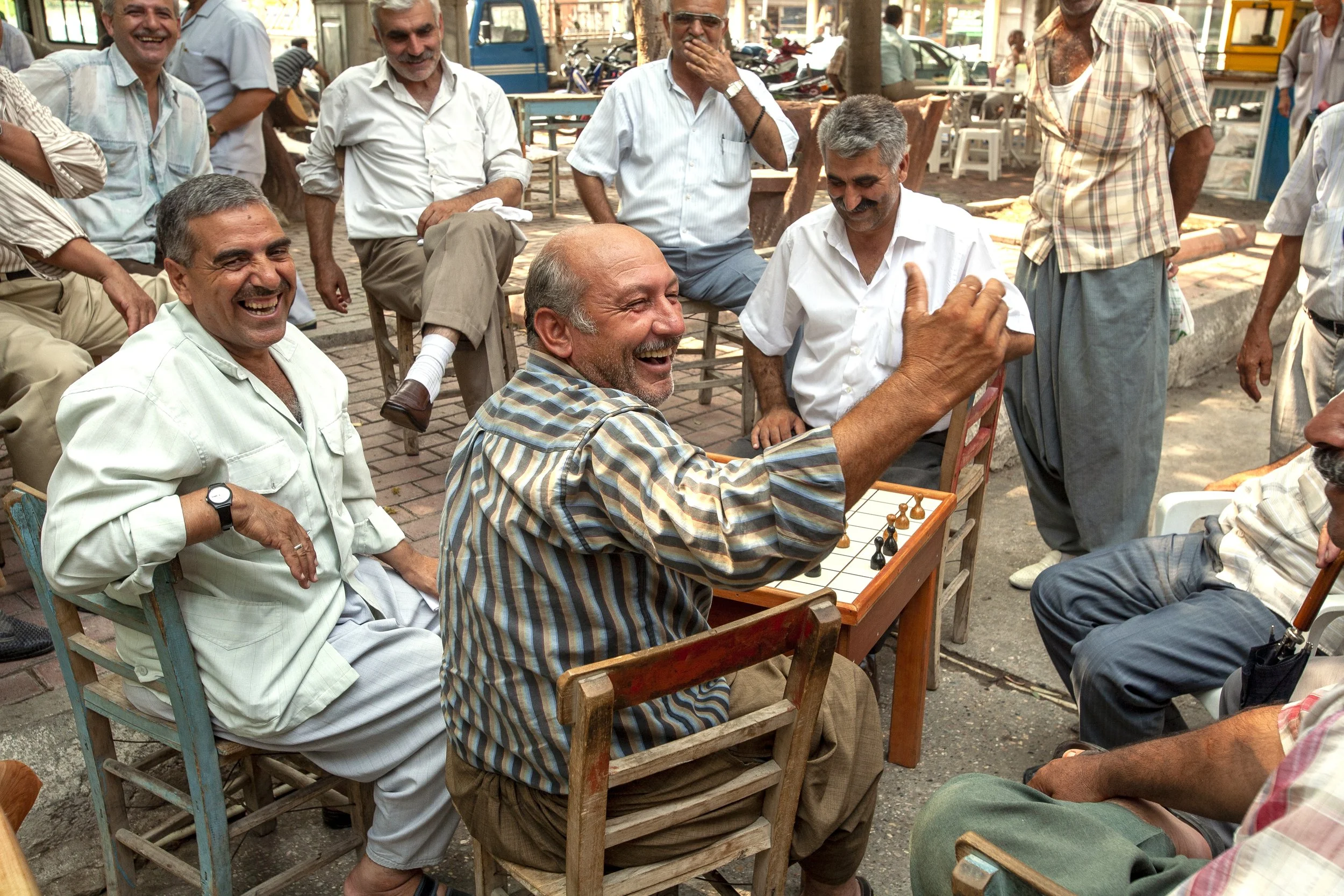 A group of men sitting and standing outdoors on a sidewalk, enjoying a conversation and laughter. One man is in the foreground seated at a table, gesturing with his hand, while others are smiling and watching, with some seated and some standing around. The setting appears casual, with motorcycles and stores in the background.