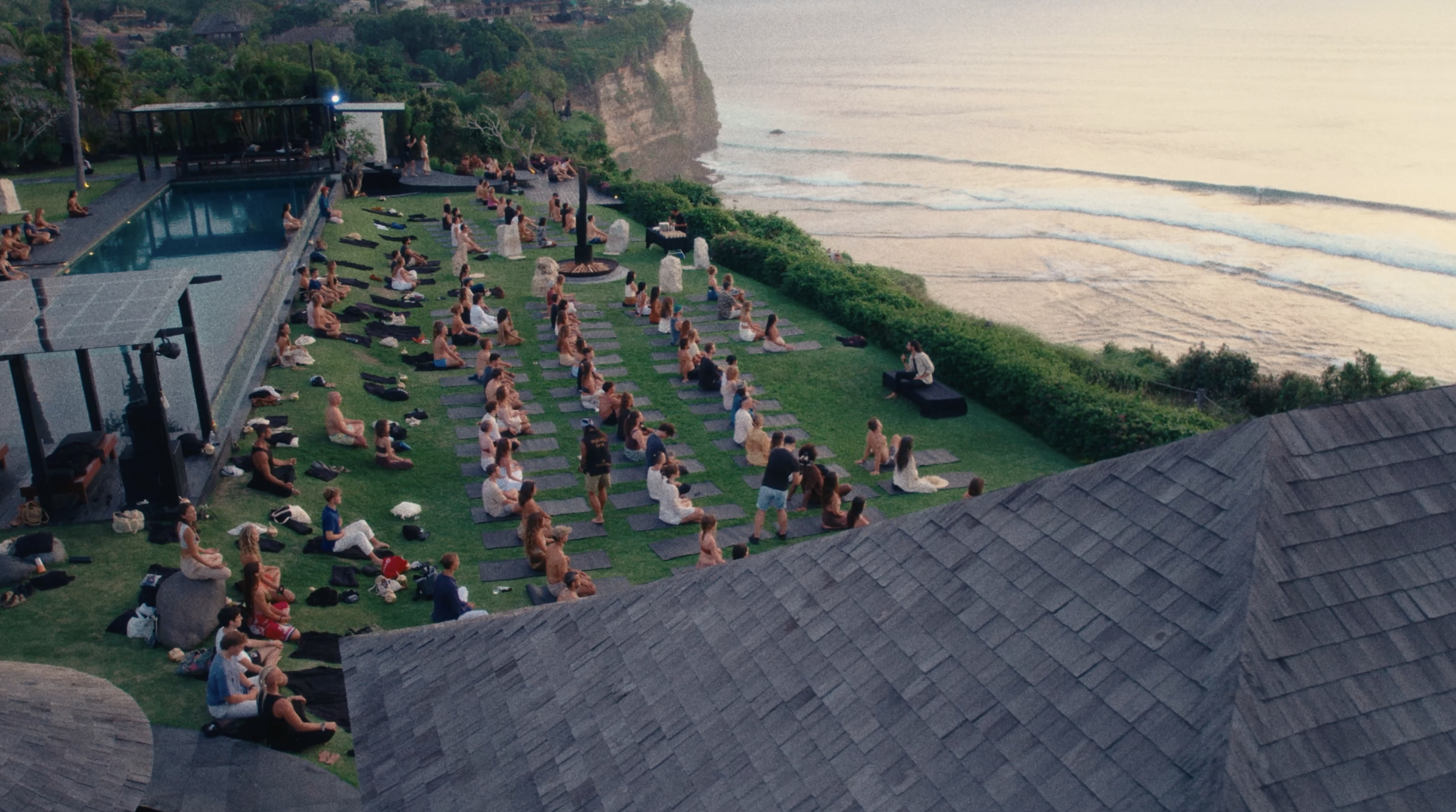 People practicing yoga on a lawn near the ocean with a pool and cliffs in the background at sunset.