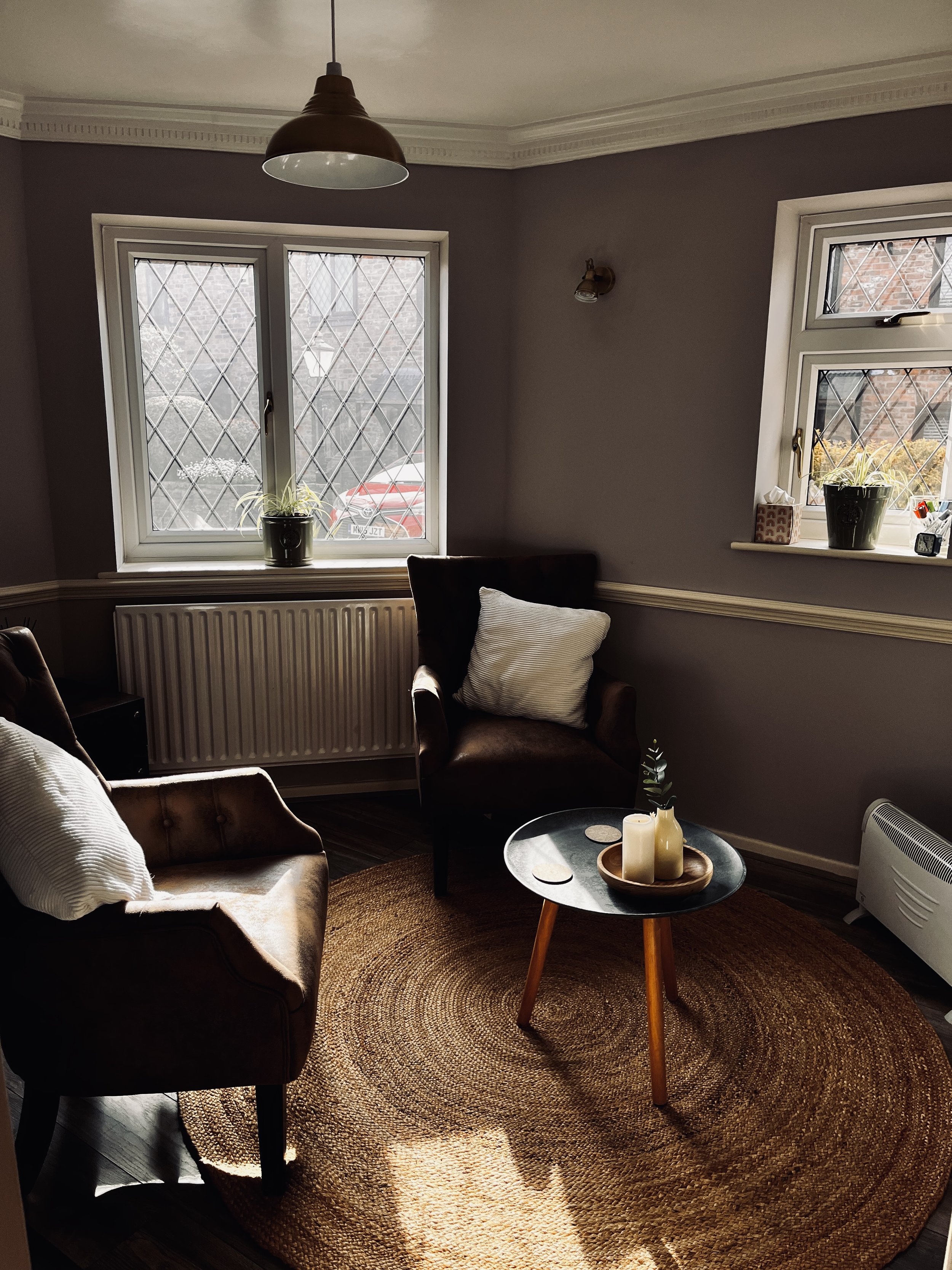 A cozy living room with a round woven rug on dark hardwood floor, two brown armchairs, and a small black round coffee table with candles and a small plant. Two windows with diamond lattice panes, one larger and one smaller, with white trim and window