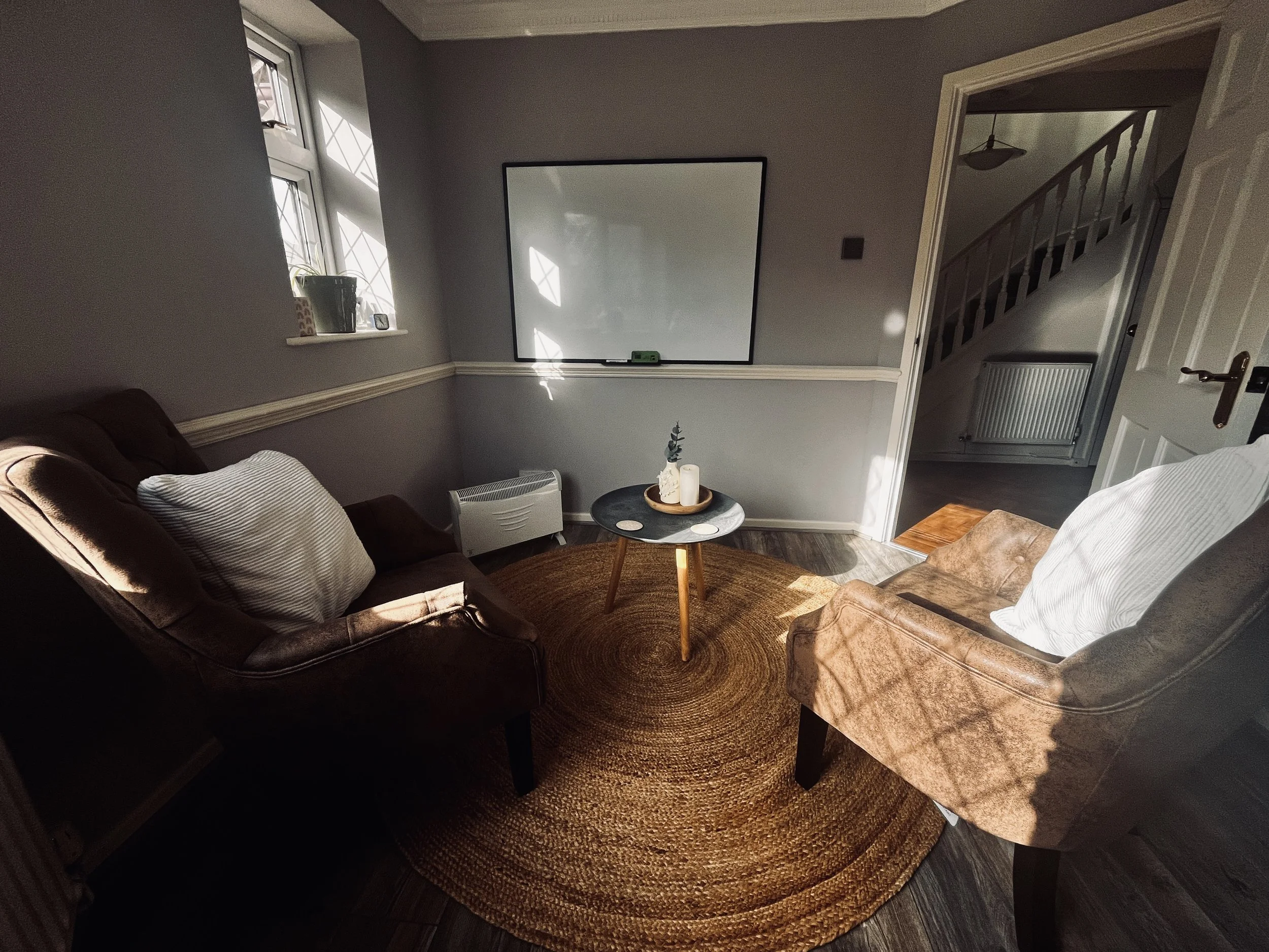 A cozy living room with two armchairs, one brown and one tan, positioned around a small round black coffee table with candles and a small plant, a white radiator under the window, and a whiteboard mounted on the wall.