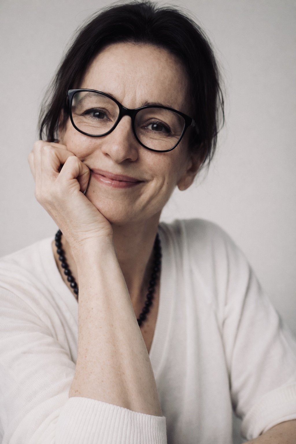 A woman with short dark hair, glasses, and a gentle smile, resting her chin on her hand.