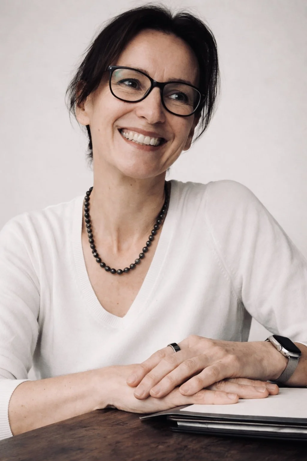 A woman with short dark hair wearing glasses, a white V-neck sweater, a beaded necklace, a smartwatch, and a ring on her finger, sitting at a dark wooden table with her hands folded and smiling.