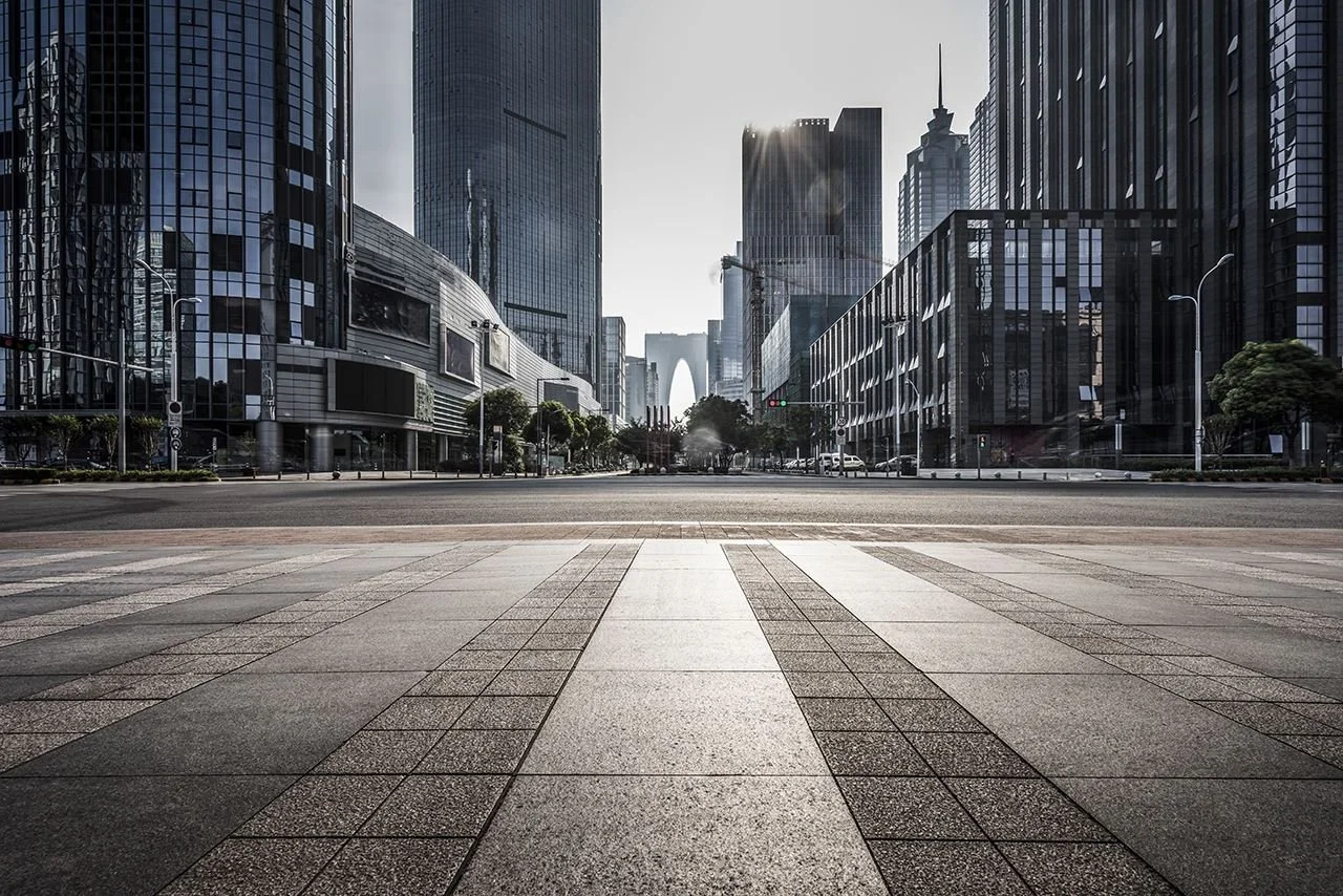Empty city street with modern skyscrapers and office buildings in the background, sunlight shining over the horizon, and a tiled crosswalk in the foreground.