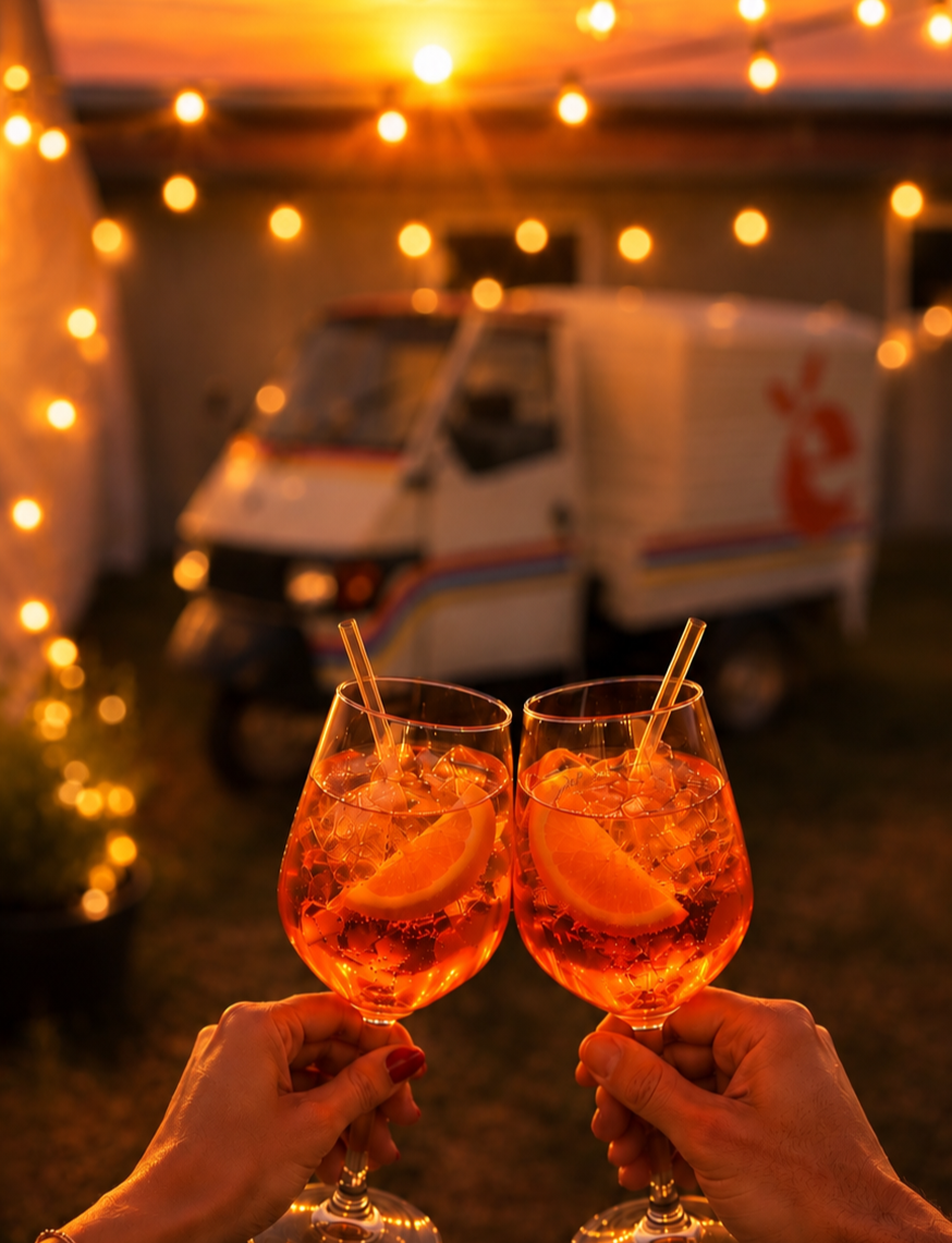 Zwei Menschen stoßen mit Aperol Spritz an, im Hintergrund eine beleuchtete Vintage-Wohnmobil bei Sonnenuntergang.