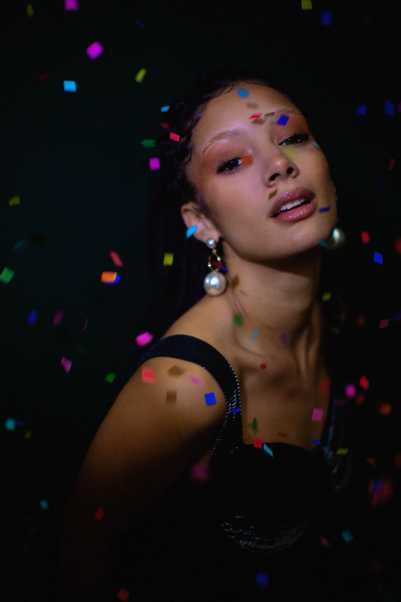 Une femme portant de grandes boucles d'oreilles en perles, entourée de confettis colorés dans un fond noir, avec une expression confiante et sophistiquée.
