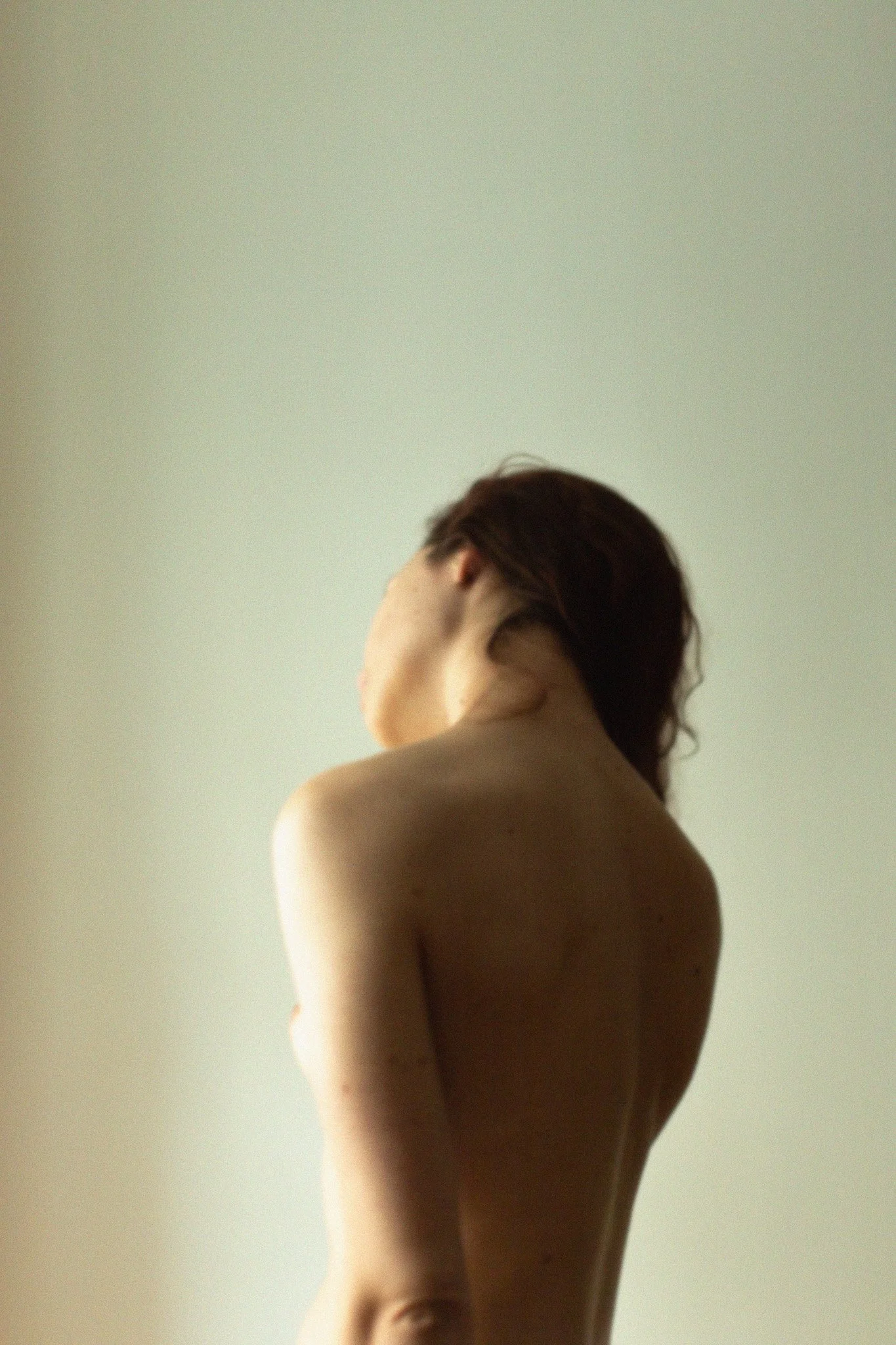 Un portrait artistique d'une femme nue vue de dos, avec des cheveux courts foncés, contre un mur clair.