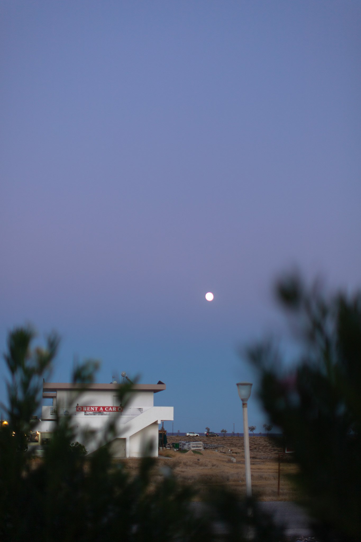 Vue d'un ciel crépusculaire avec la lune visible, vue partielle d'un bâtiment blanc avec un panneau 'Location de voitures', lampadaire dans un paysage désertique.