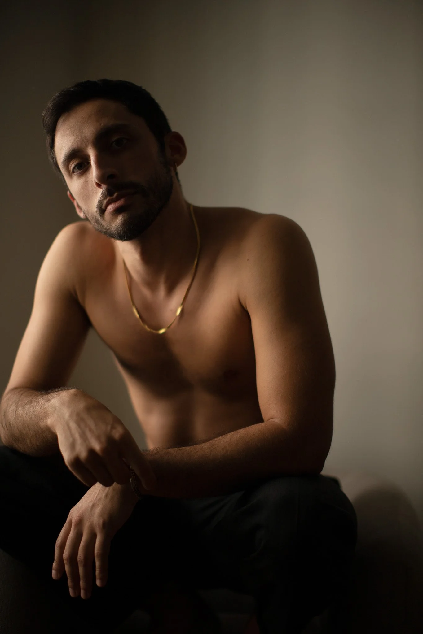 Un homme nu, assis, avec un collier en or, regardant vers l'objectif dans une pièce à l'éclairage tamisé.