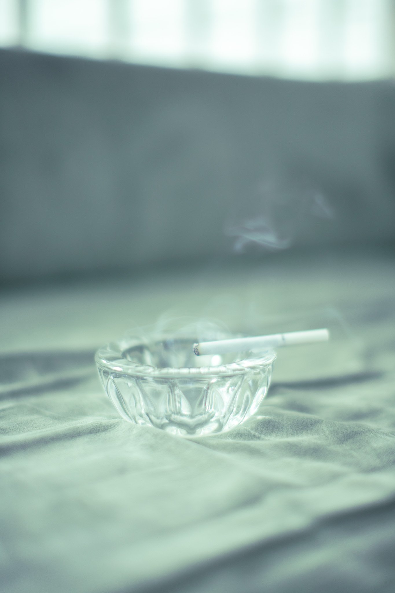 Cendrier en verre avec une cigarette à moitié consumée, placé sur un tissu clair, avec une lumière douce et un fond flou.