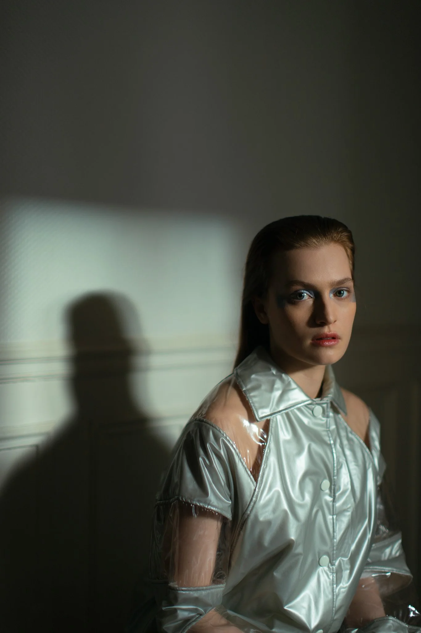 Jeune femme avec maquillage audacieux portant une veste en plastique argentée, assise ou debout dans un espace sombre avec un jeu d'ombre et de lumière.