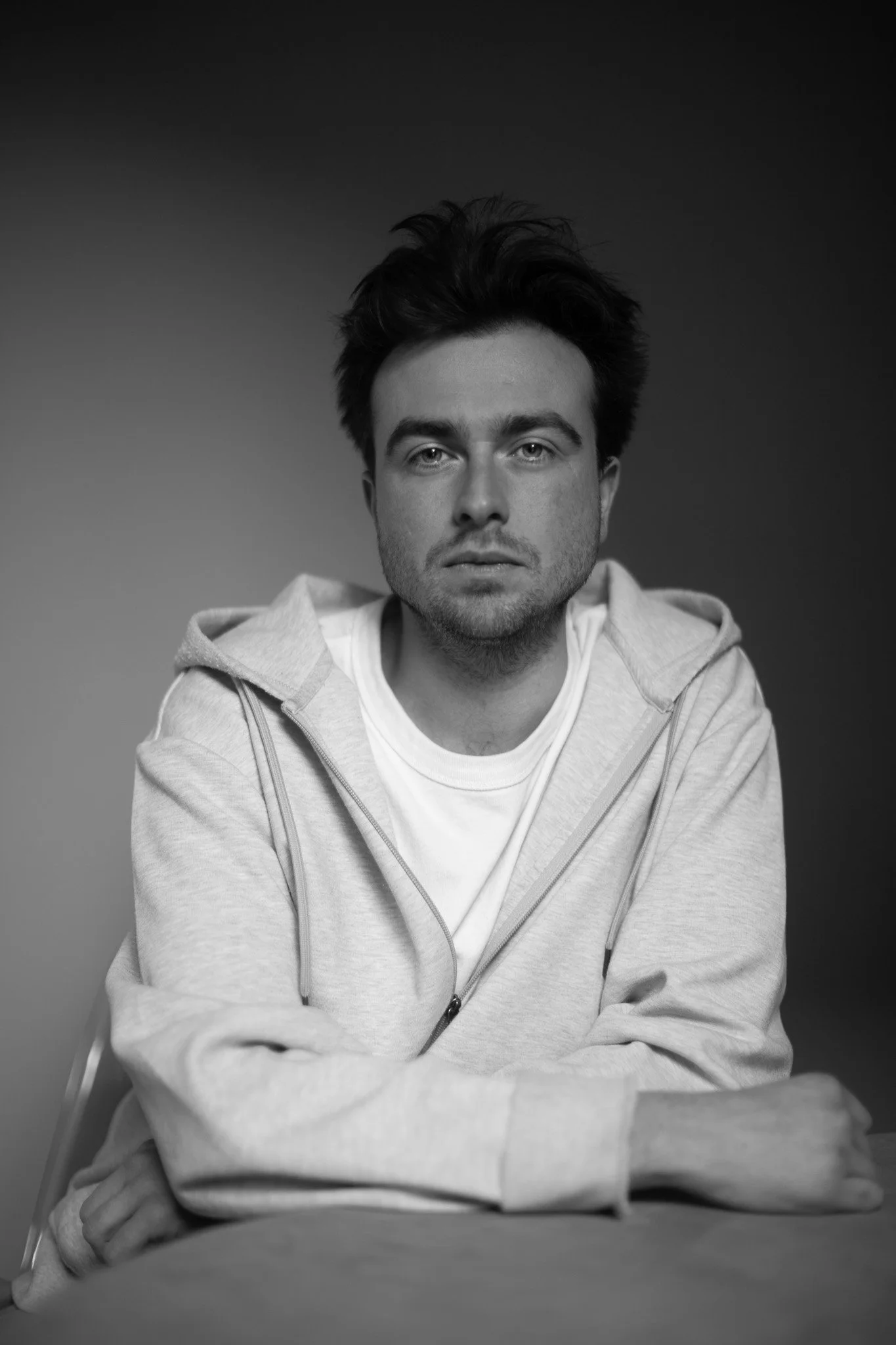 Un portrait en noir et blanc d'un jeune homme assis, portant un sweat à capuche et un t-shirt blanc, avec une expression neutre. Arrière-plan sombre.