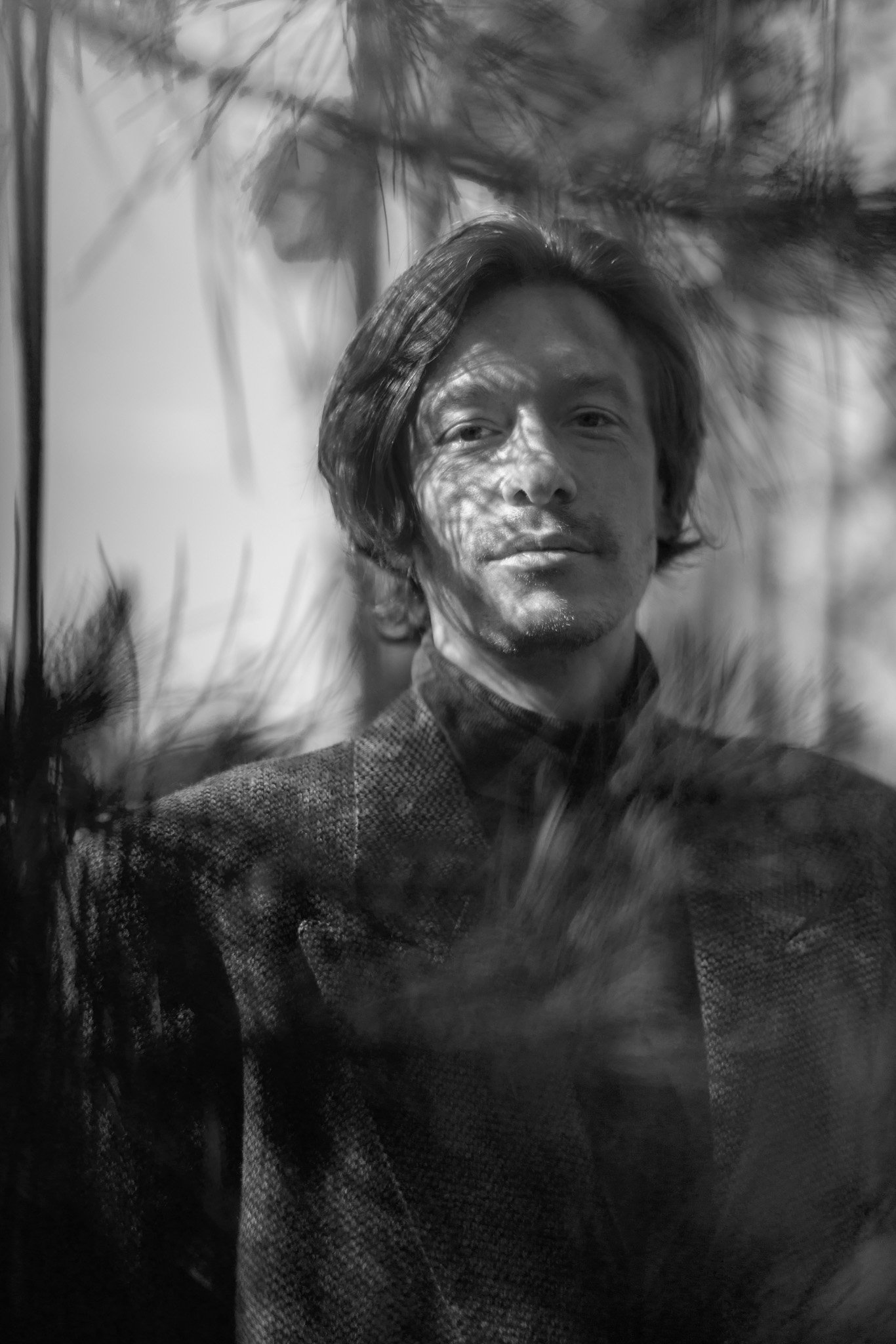 Portrait en noir et blanc d'un homme avec des cheveux longs, portant un costume sombre, avec des reflets de lumière et d'ombres sur son visage.
