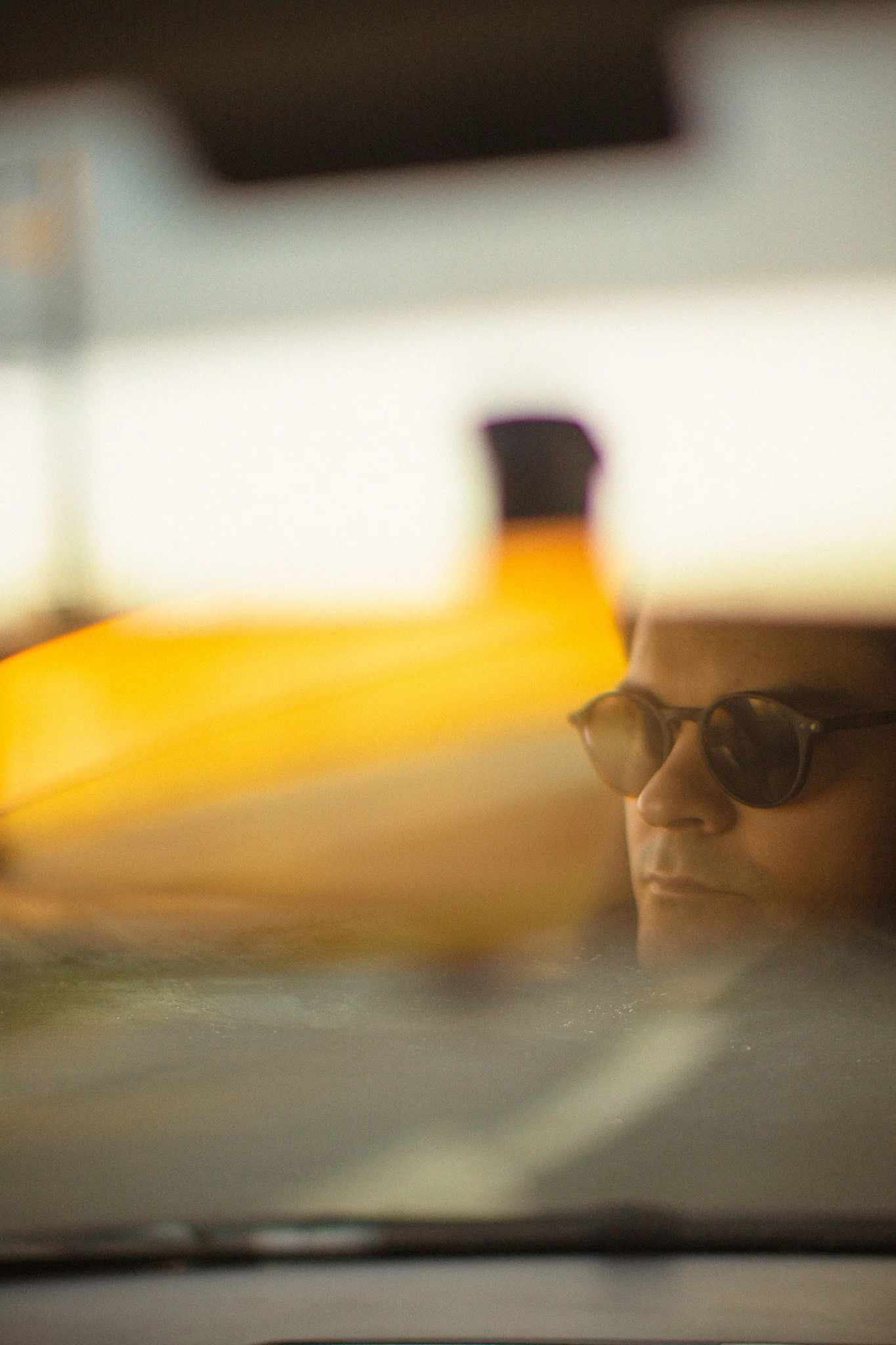 Un homme portant un chapeau et des lunettes de soleil regarde vers la droite, avec un fond flou et un objet jaune à l'avant.