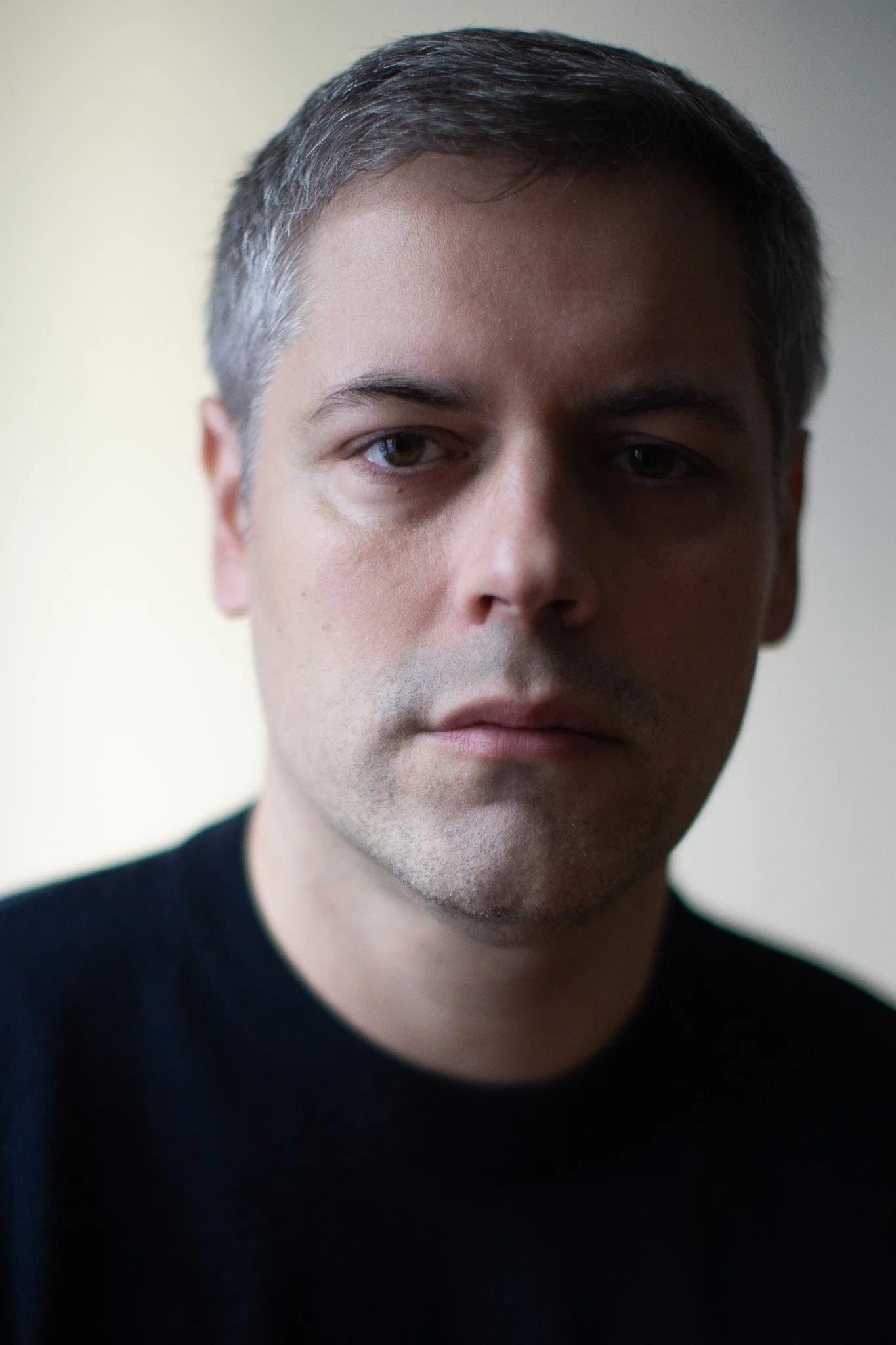 Portrait d'un homme avec des cheveux gris et un t-shirt noir, regardant vers l'objectif, dans une lumière douce et neutre.