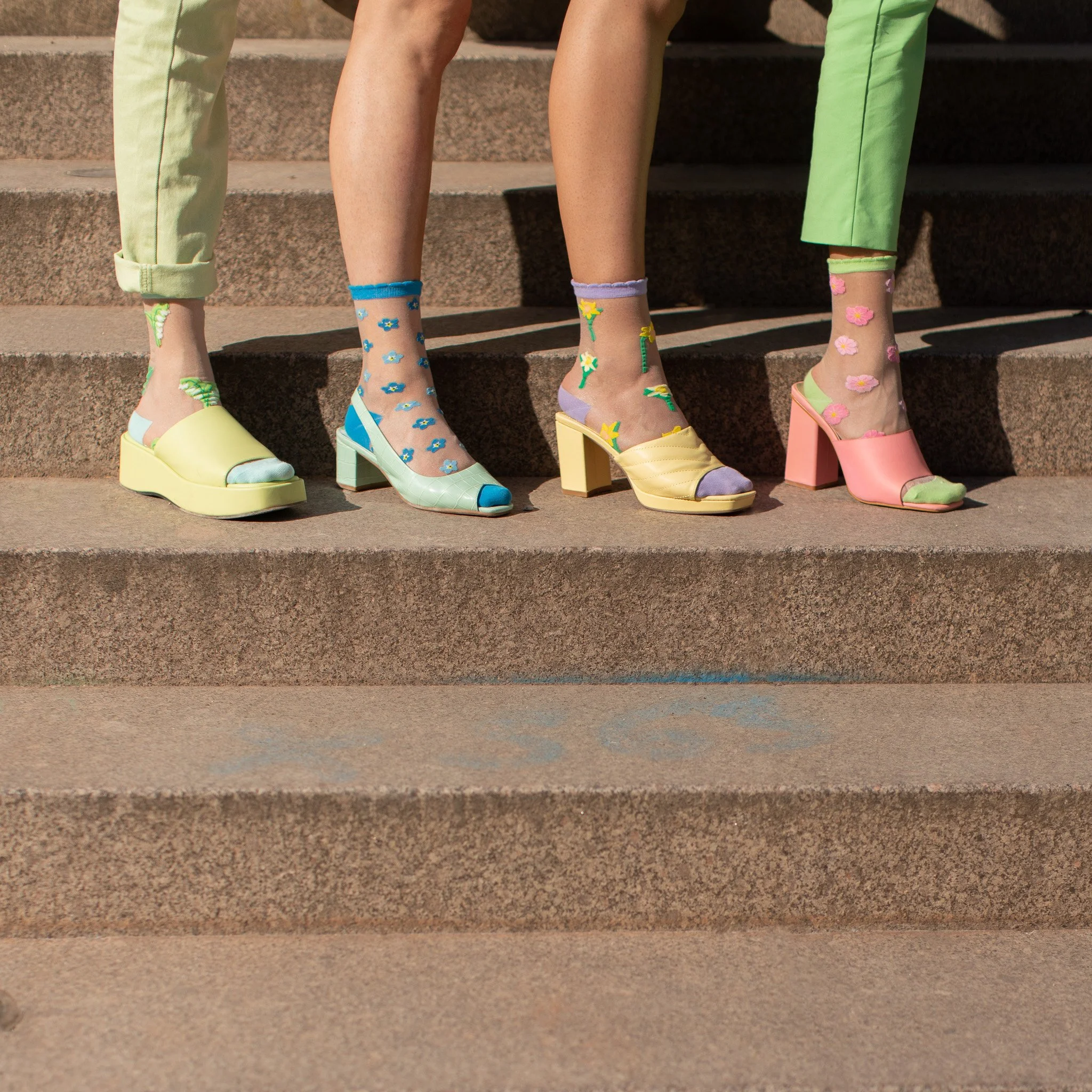 Gros plan des jambes et chaussures de trois personnes portant des chaussettes colorées, des sandales à talons hauts et des pantalons de couleurs vives, assises sur des marches en pierre.