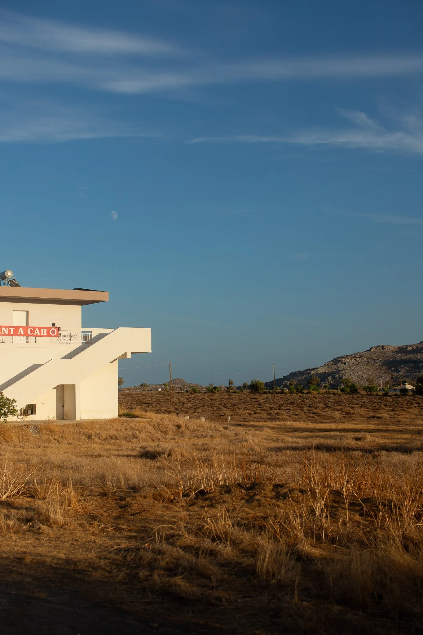 Un bâtiment blanc avec un escalier extérieur dans un paysage désertique, sous un ciel bleu avec des nuages et la lune visible.