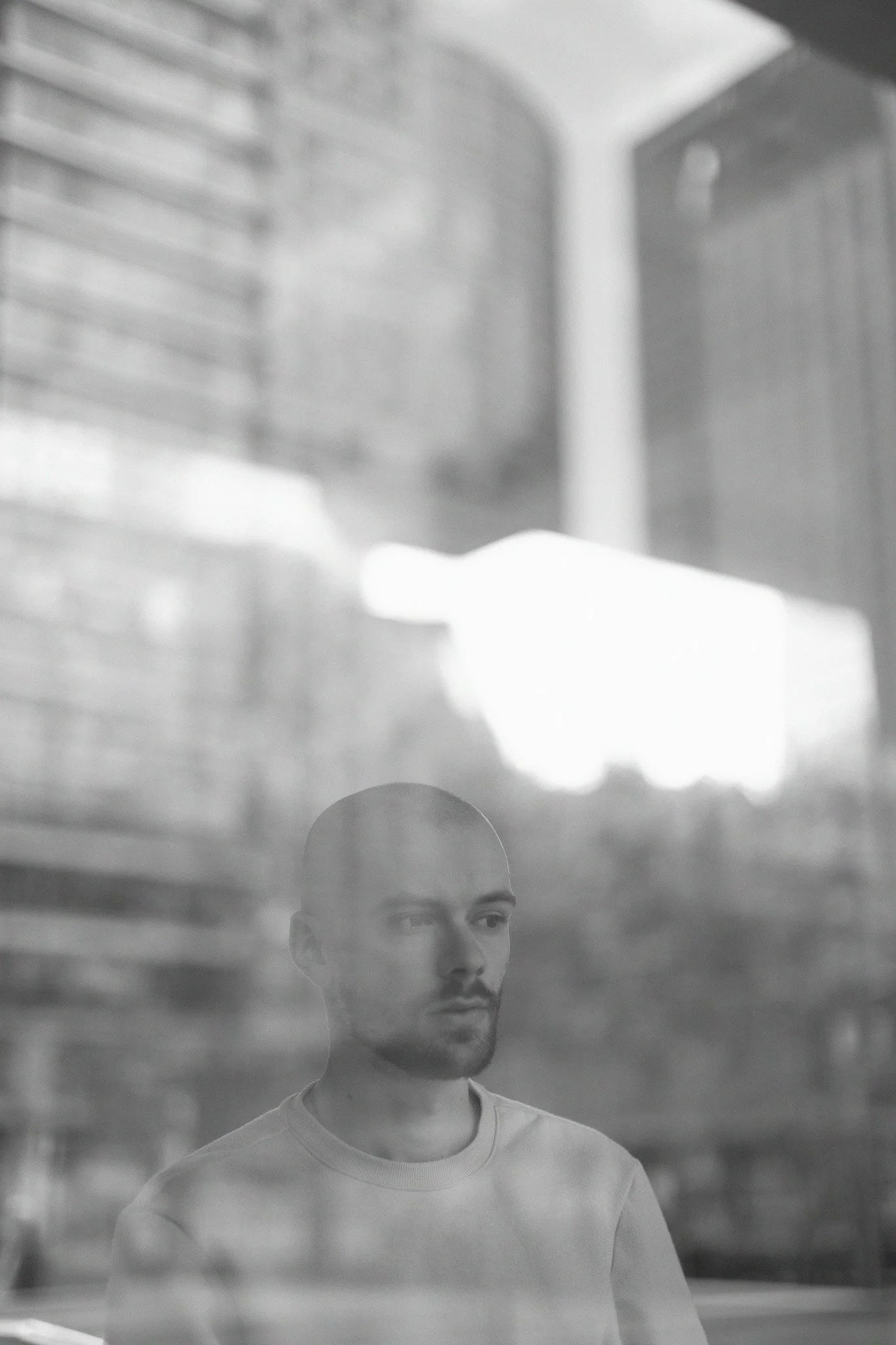 Un homme regarde à travers une vitre, reflet de son visage visible sur la surface transparente, avec un arrière-plan flou de bâtiments modernes.